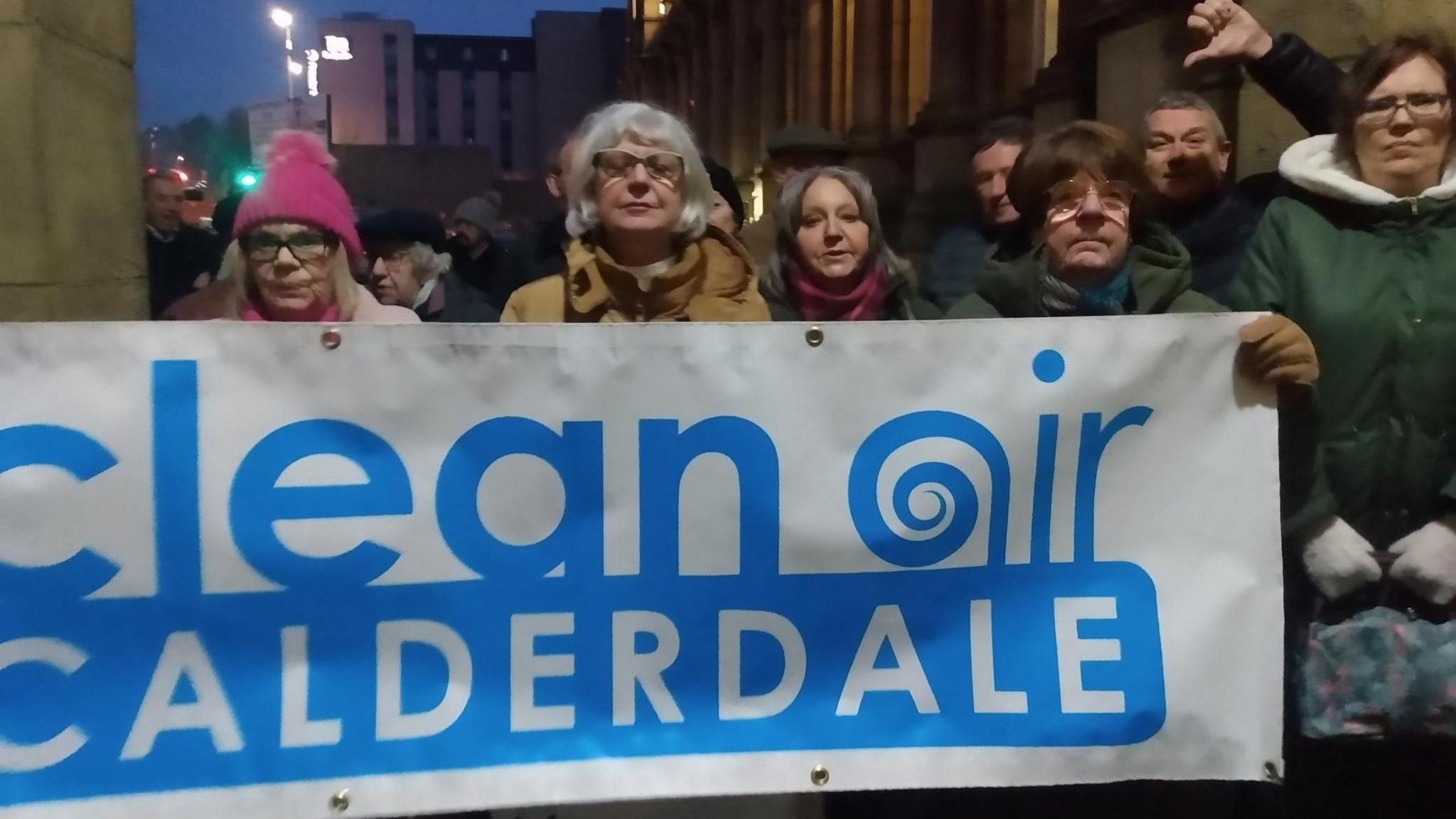 About eight people in the foreground, with others blurred in the background, holding a banner reading "clear air calderdale". It is nighttime. A man gives a thumbs down in the background.
