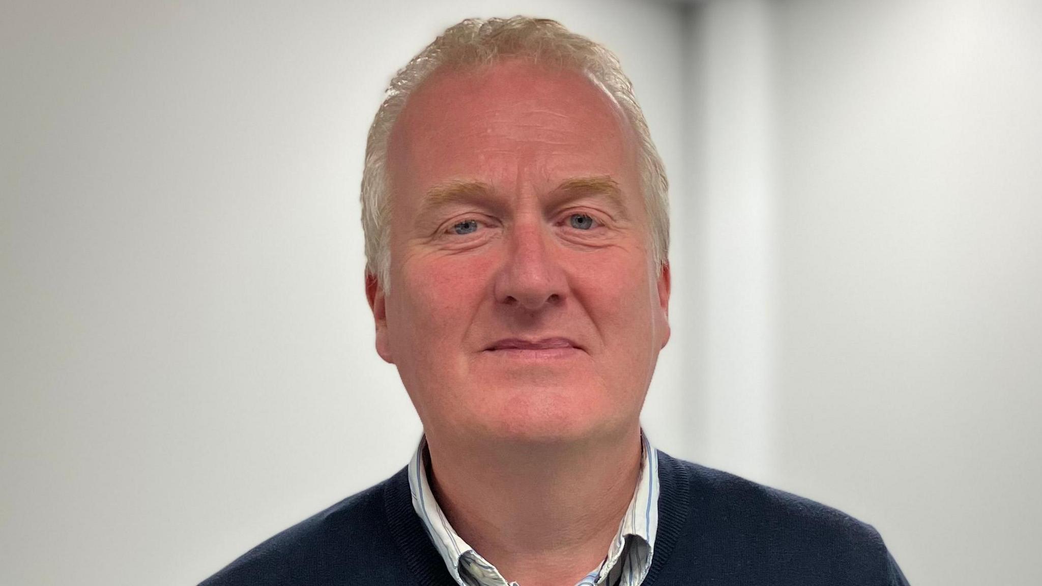 Eugene Reid is wearing a navy jumper with a stripped shirt underneath. He has grey hair and is standing in front of a white wall.