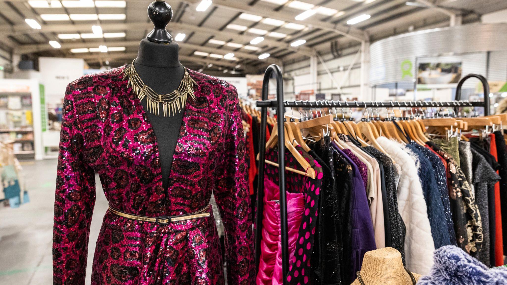 A mannequin and a rack of clothes in a charity shop. The mannequin is dressed in a red leopard print dress and a spiky layered necklace. Various types of clothes are on clothes hangers on the rack.