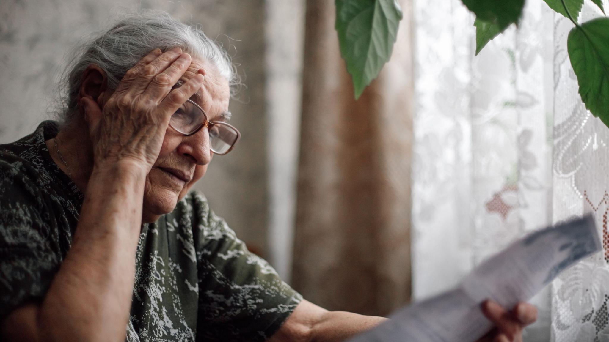 An elderly, bespectacled woman holds her head in one hand and a paper bill in the other