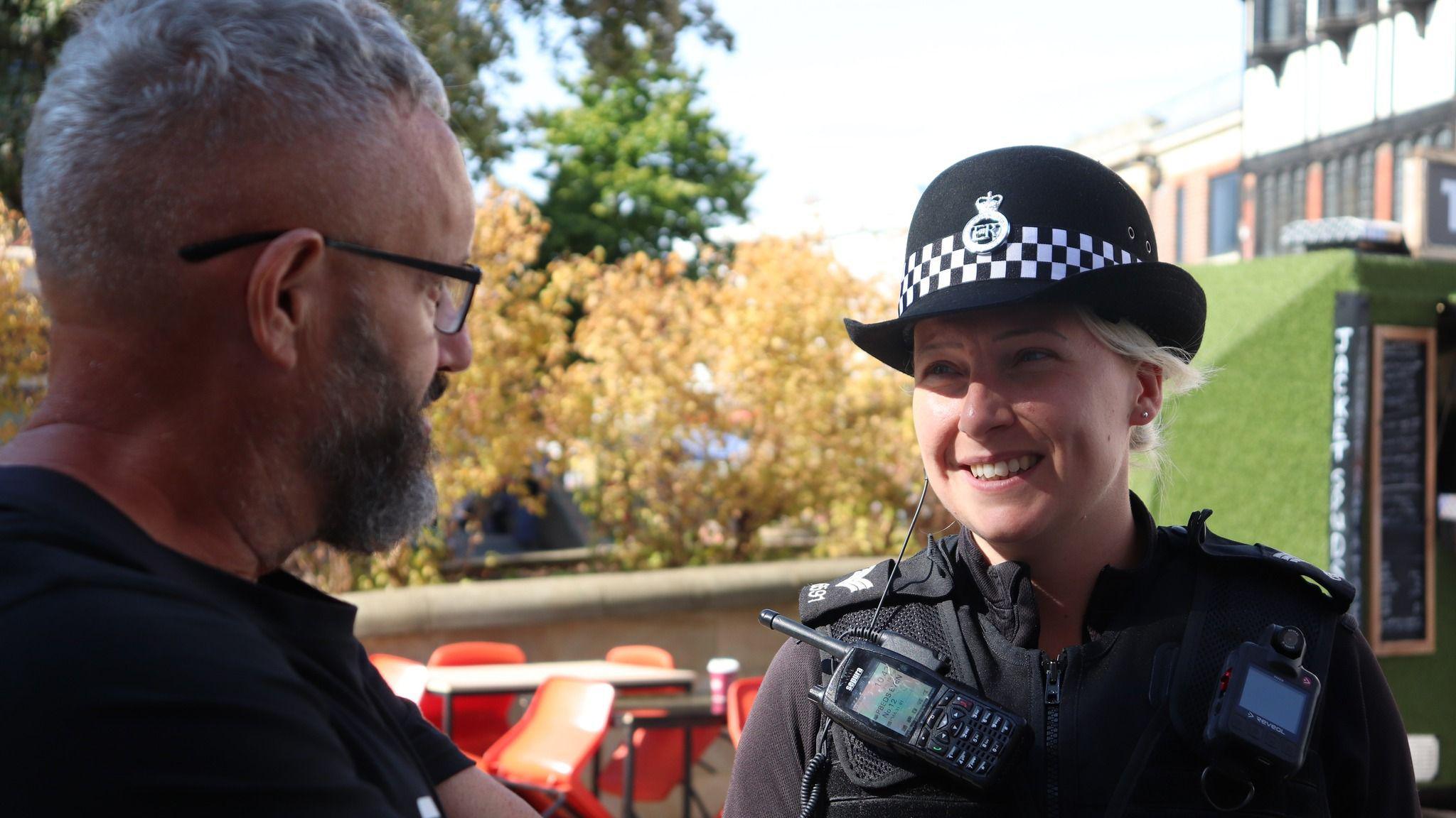 A female PC, in full black police uniform, with a hat on her head, with blonde hair tied back, talking to a man. She is smiling. There are table and chairs behind her. The man has only one side of his face to the camera, has short hair, has on glasses and has a beard. 