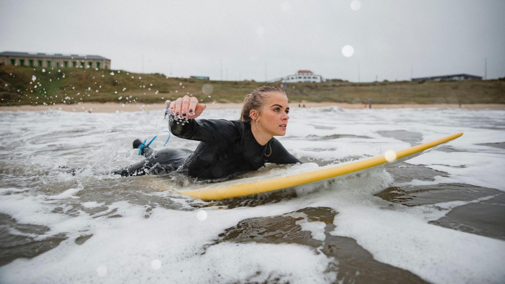 A young woman with long blonde hair lies on a yellow surfboard and paddles out on the sea. Behind her is the beach and a cliff atop of which sits several small buildings. The picture is taken at Tynemouth.