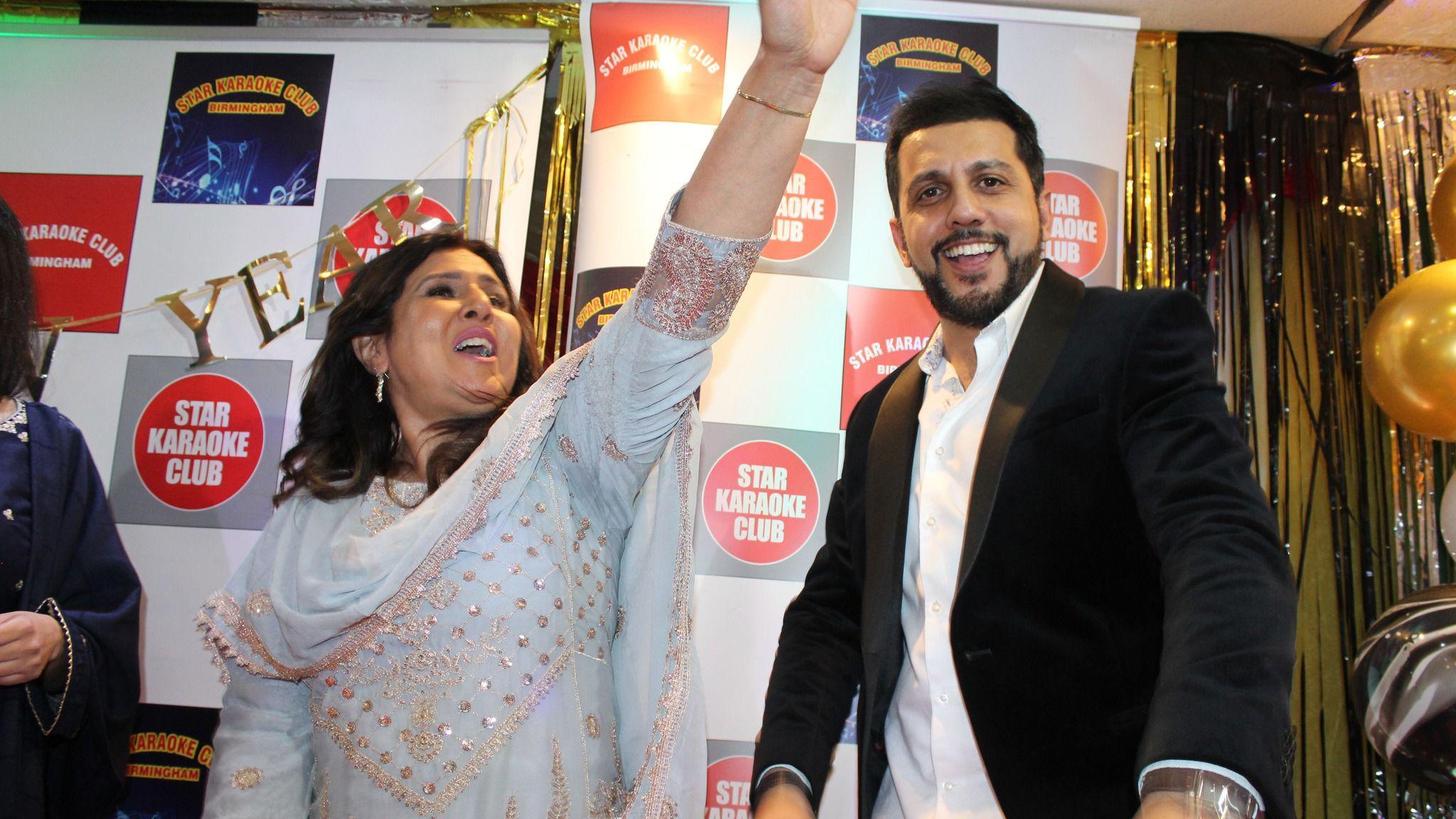 A woman is wearing a light blue dress with gold detailing, and has her arm in the air as she sings. A man next to her is wearing a black suit jacket and a white shirt, and is smiling at the camera