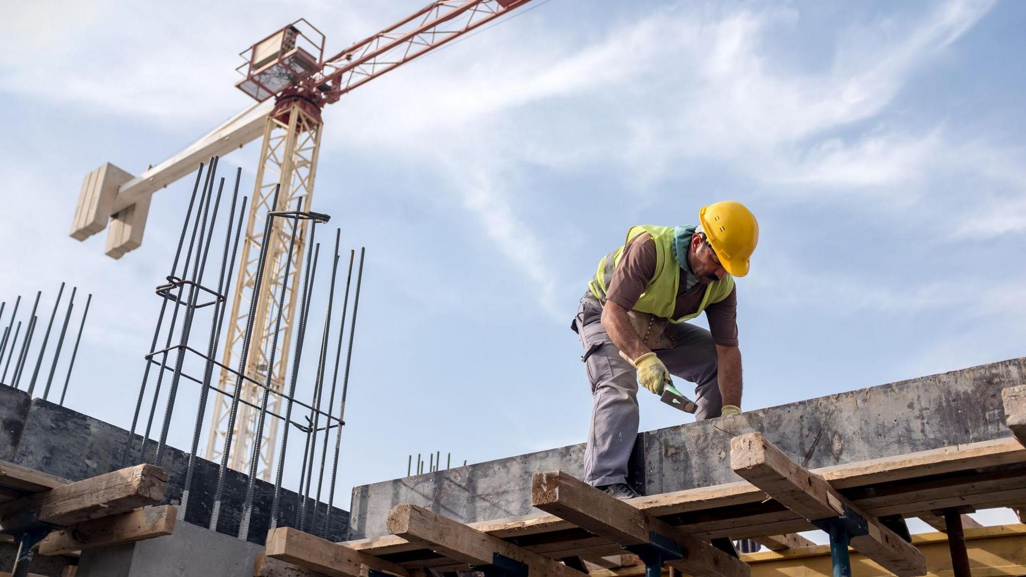 Worker on a construction site stands on a beam hammering a nail into a piece of wood