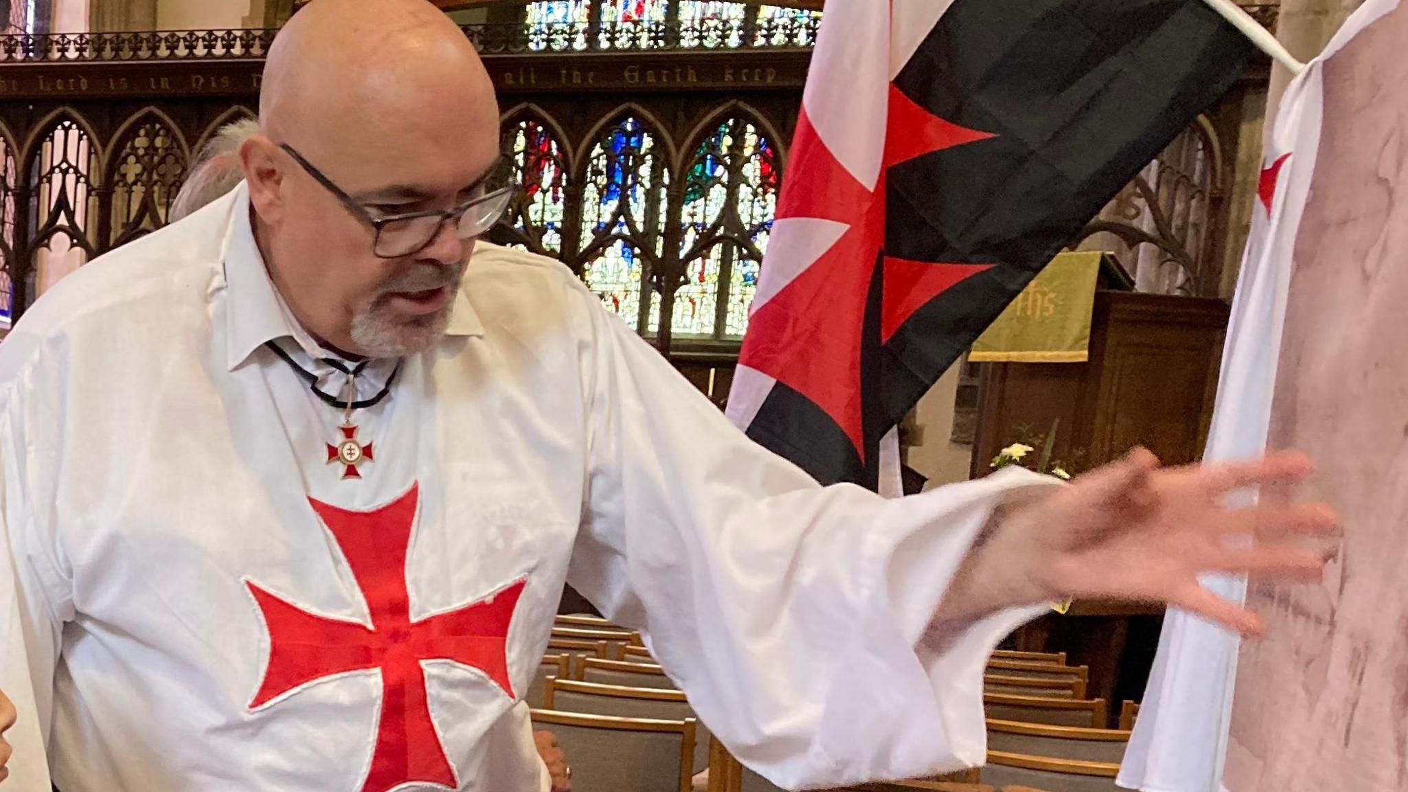 A bespectacled Brother John dressed in a white robe with a red cross on it