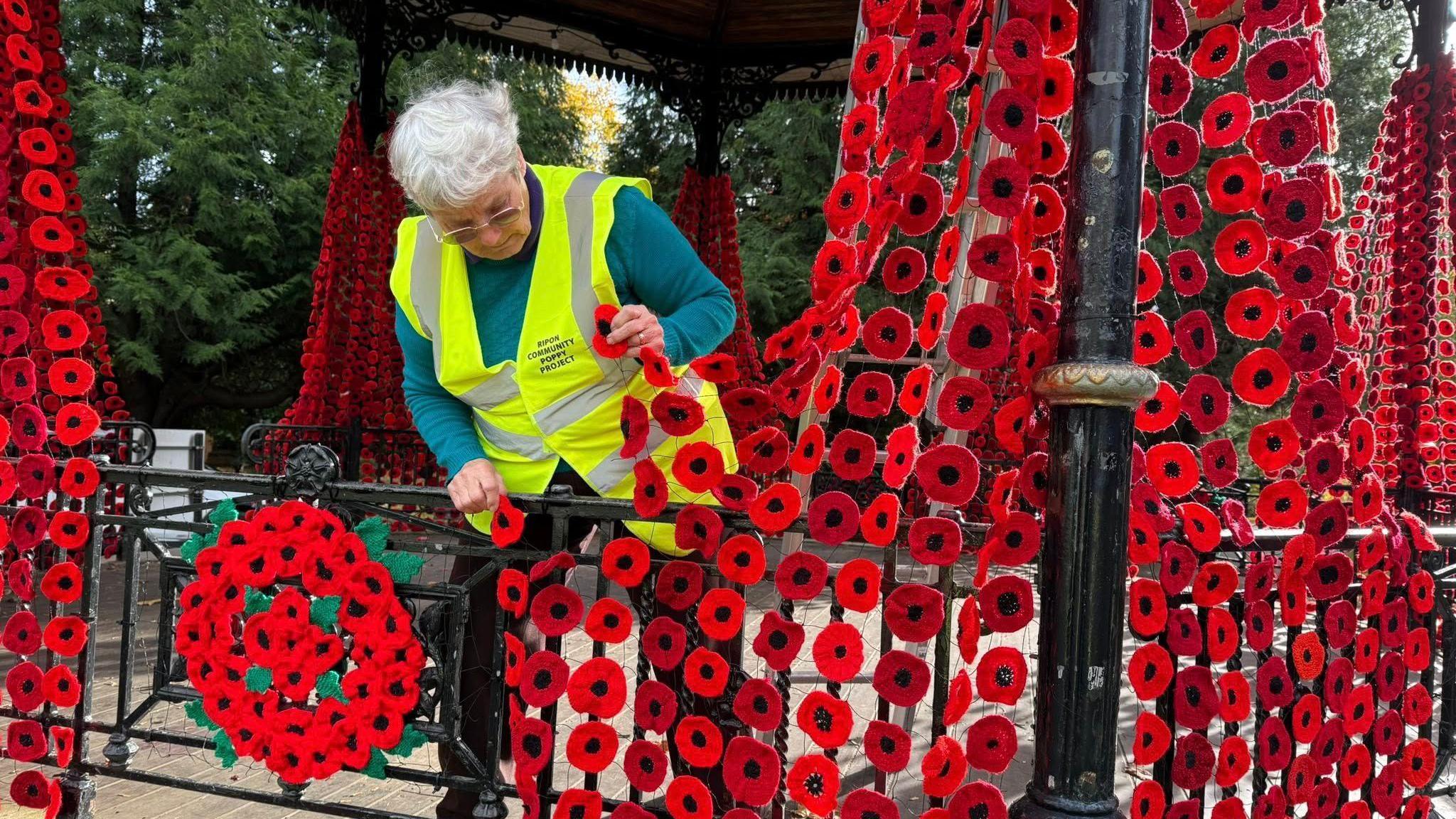 A woman wearing a bright yellow safety vest examines damage to netting displaying red poppies on a black metal bandstand in a park. The poppies hang in vertical rows and form a large circular arrangement on the railing.