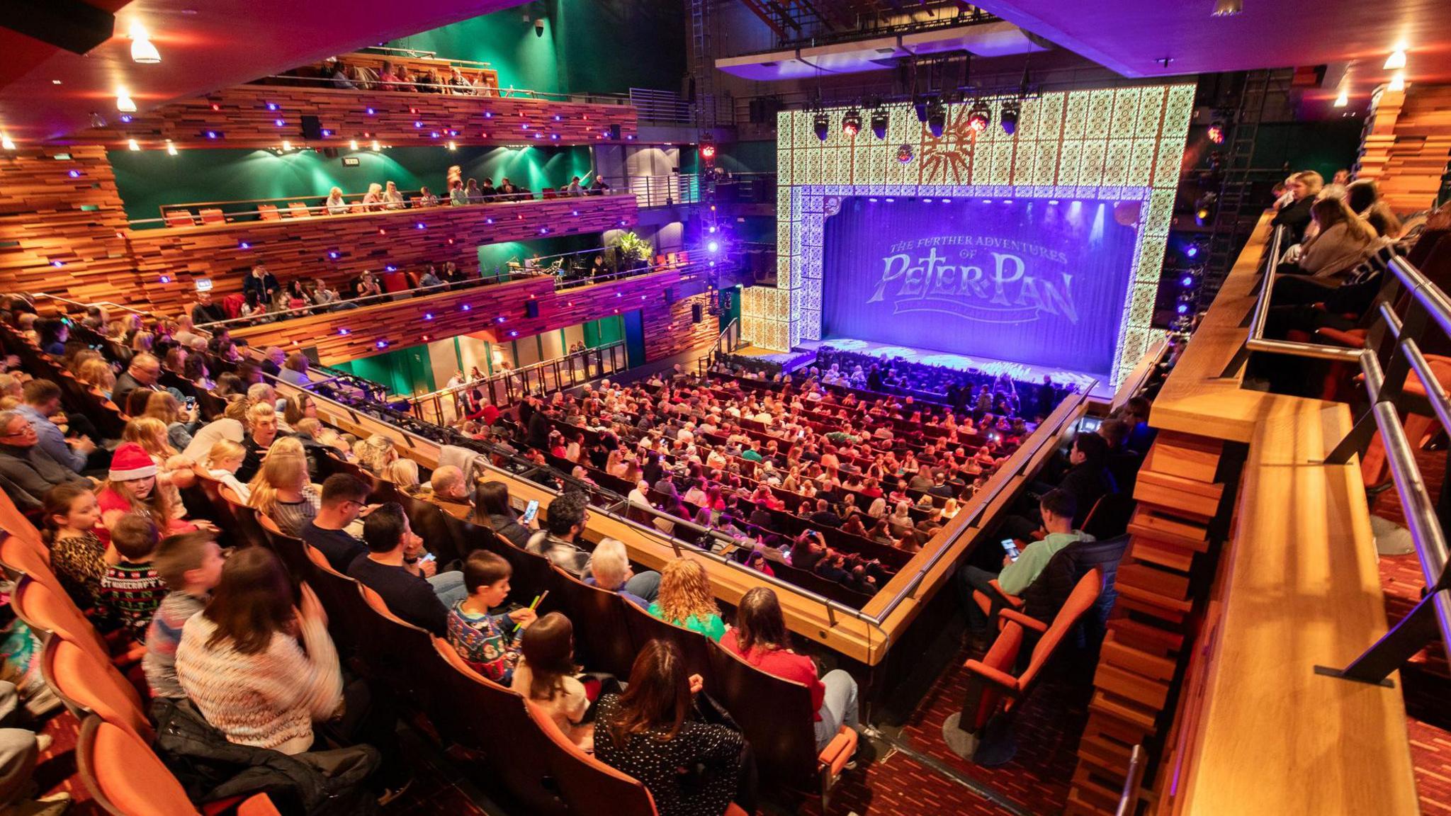 A general view of the audience inside Aylesbury Waterside Theatre. The stage says "Peter Pan" on it and is purple and gold. The seats are full with people.