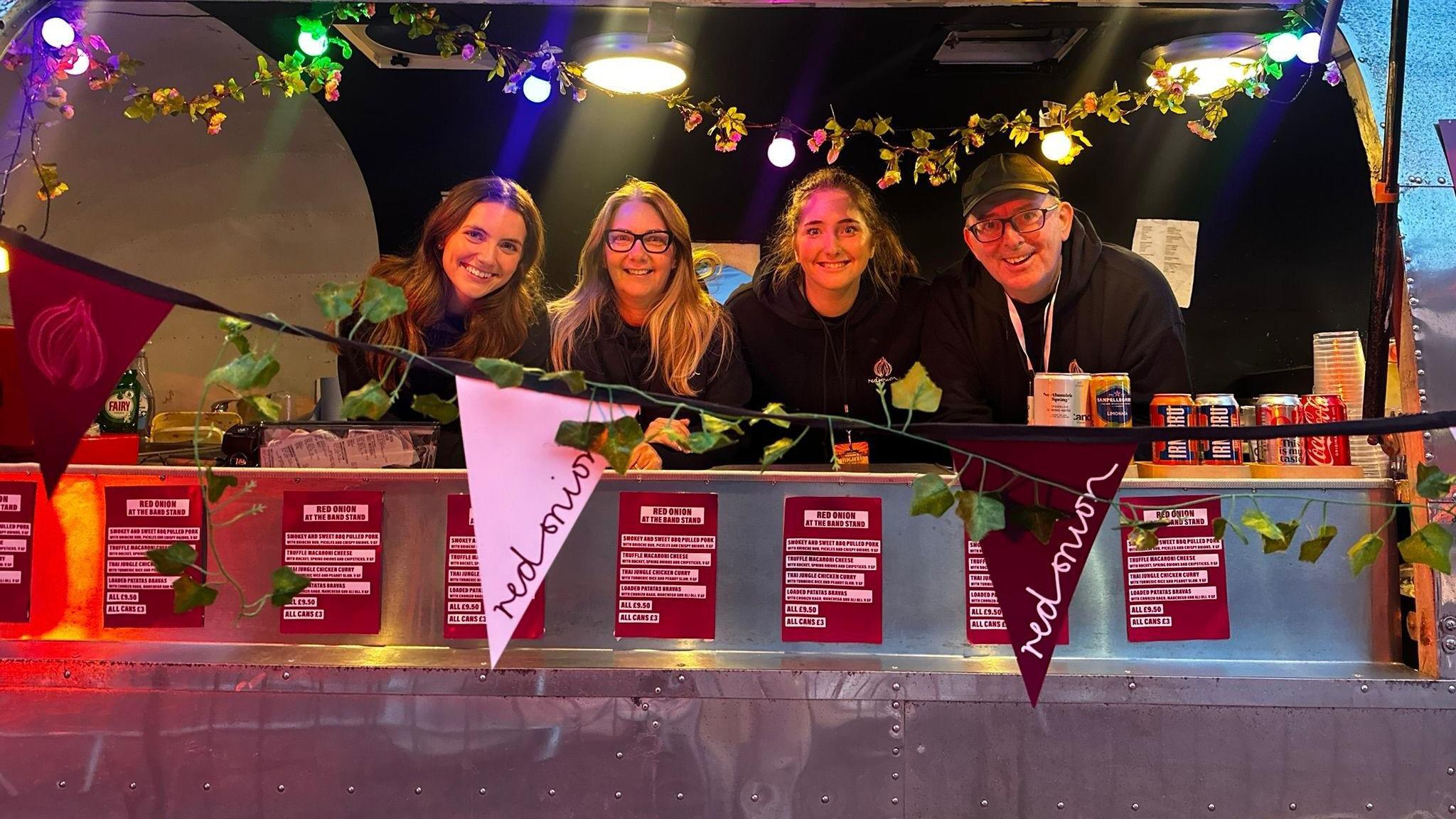 Four people standing behind the counter of a food truck at a music festival. There are three women and one man, John Quigley, who is wearing a baseballl cap. All are smiling for the camera. 