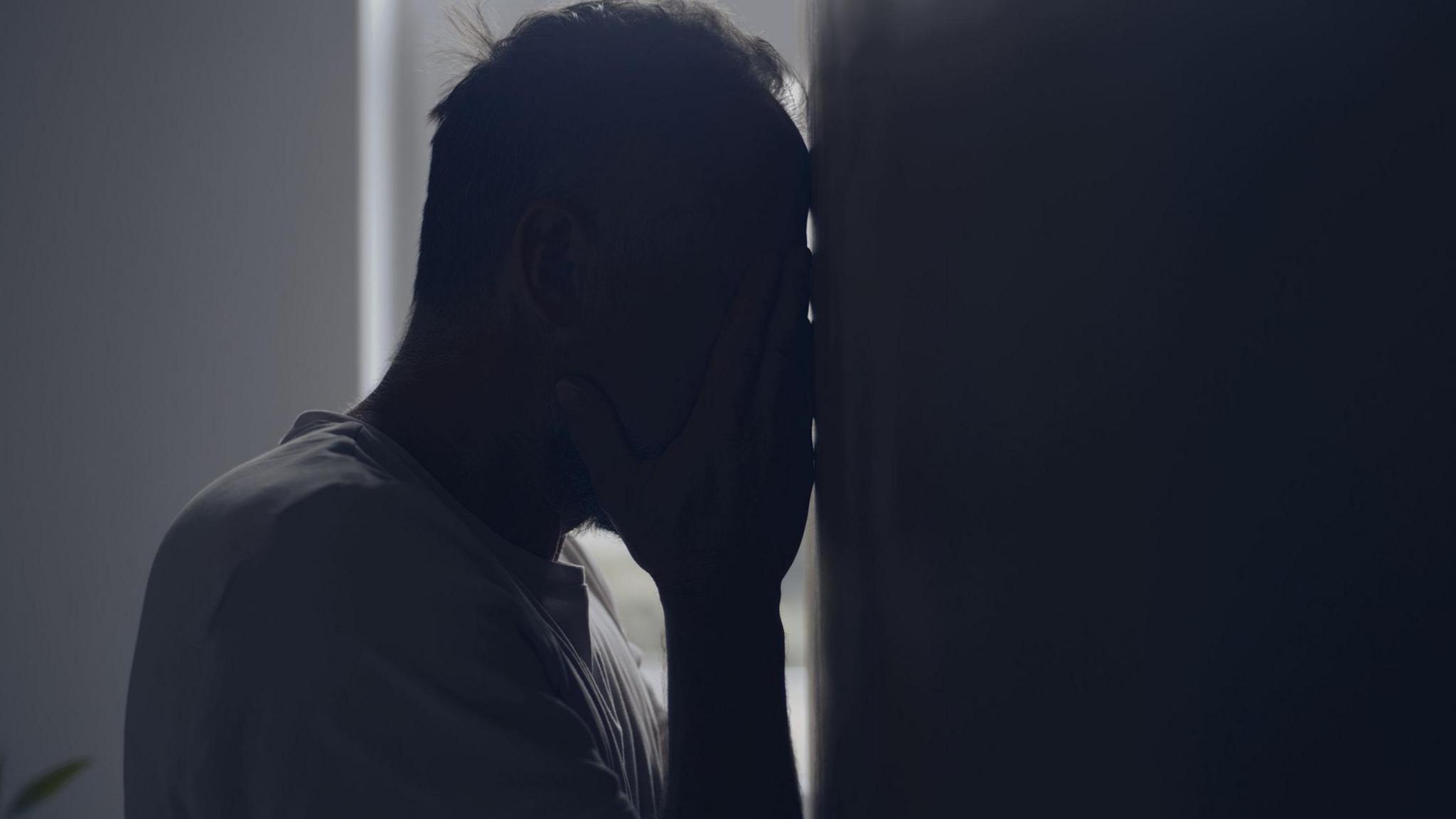 A man standing with his head against a wall covering his face with his hands.