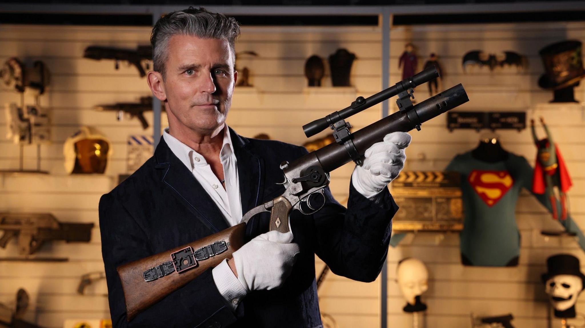 A man is holding a gun from Star Wars while stood in front of a cabinet full of film memorabilia.