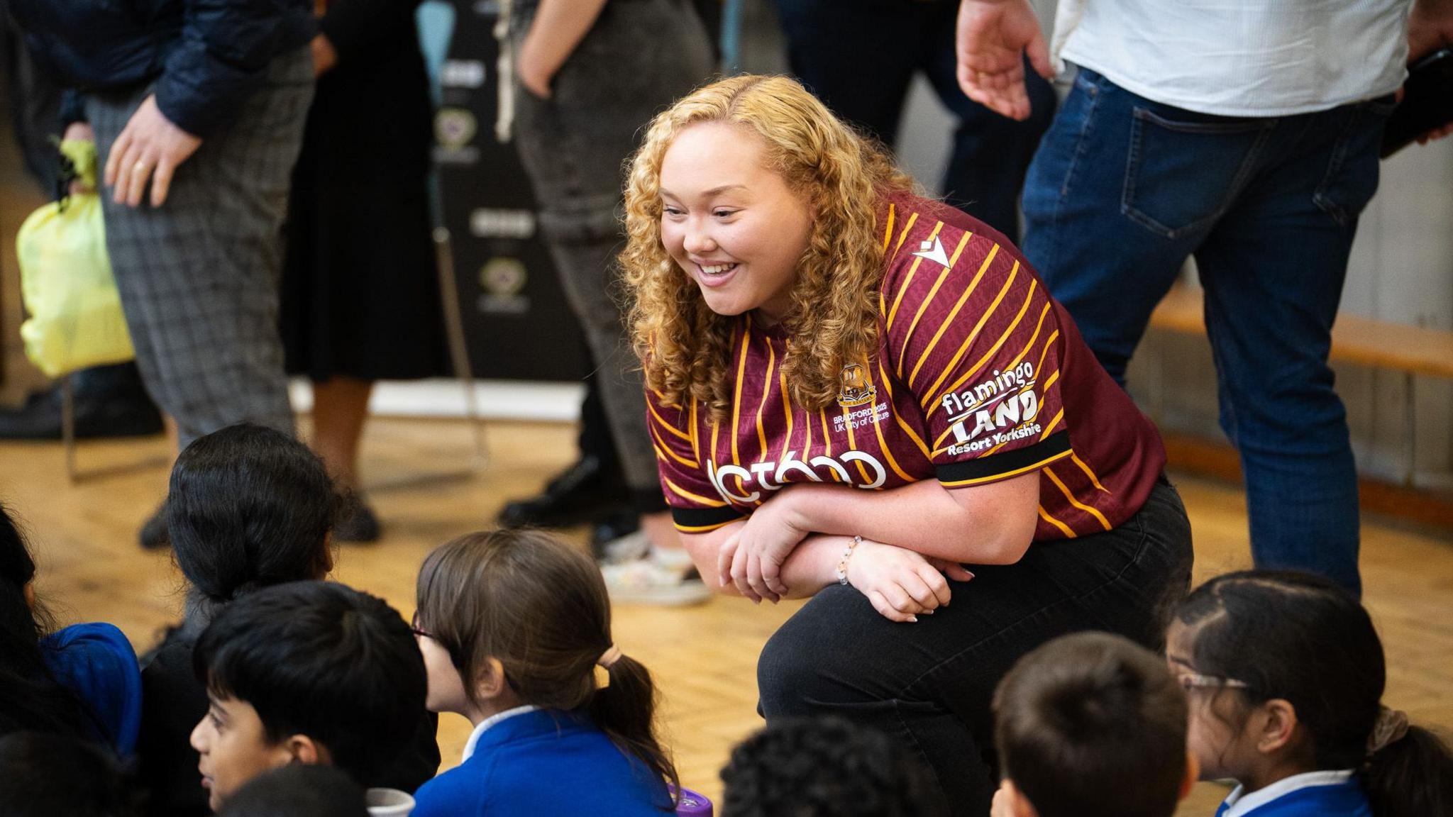 A woman with blonde curly hair and wearing a Bradford City shirt kneels down and is talking to a group of school pupils who are sat on the floor. Behind her, people are standing.