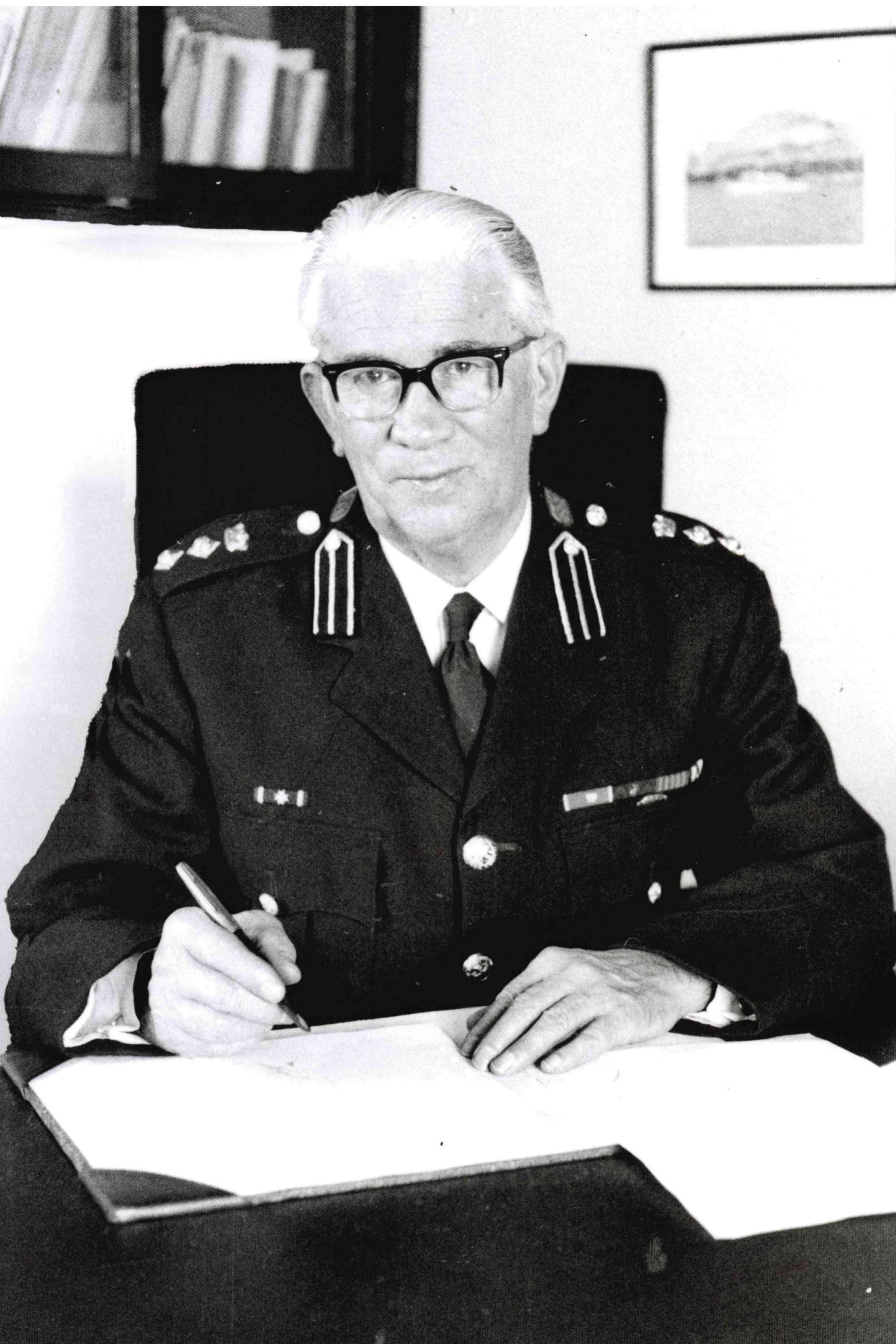 An older man with grey hair and glasses in an emergency services uniform. He sits at a desk ready to sign a book with a pen.