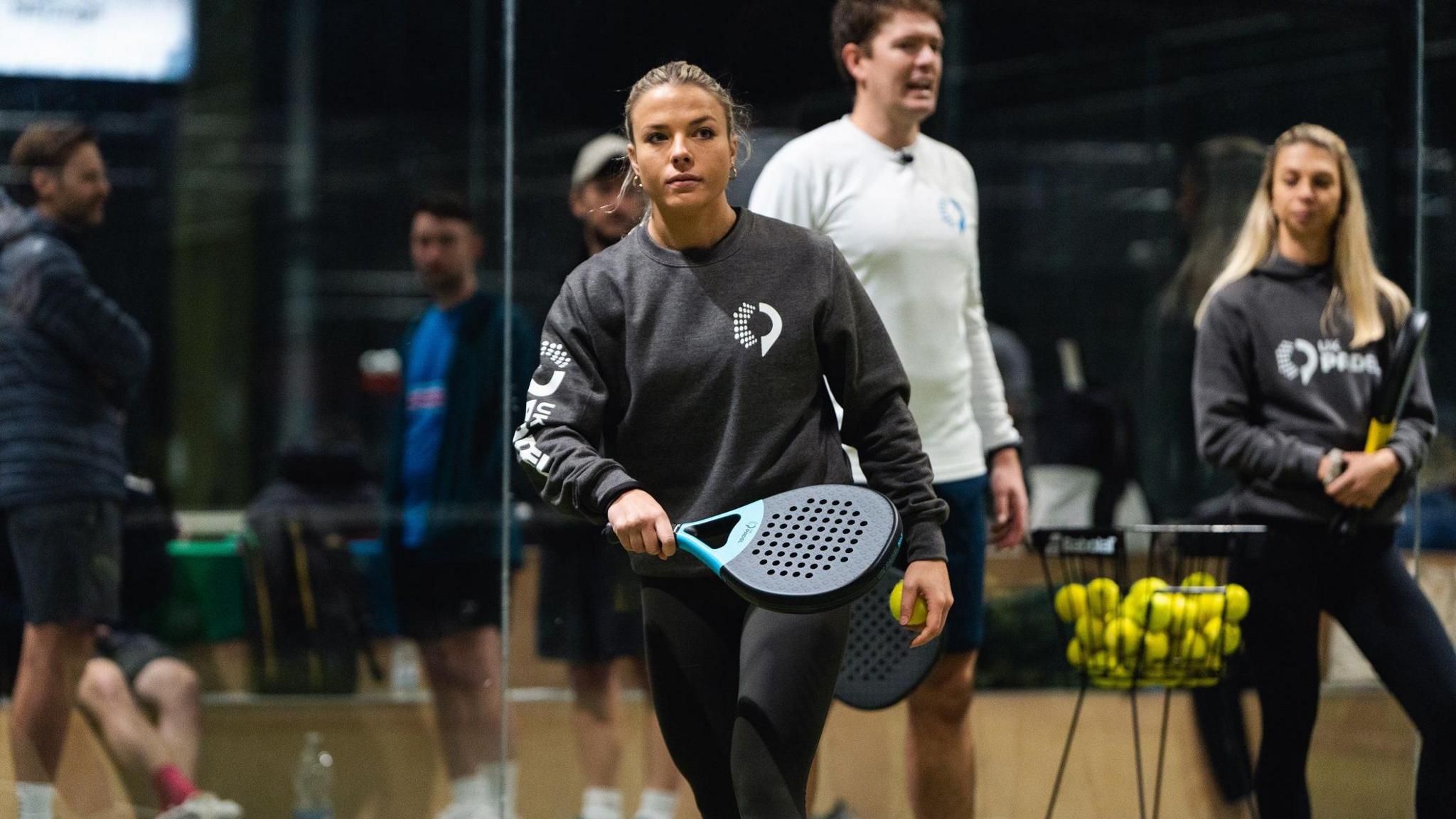 A young woman wearing all black carrying a sports racket and a ball. There is a man and a woman in the background