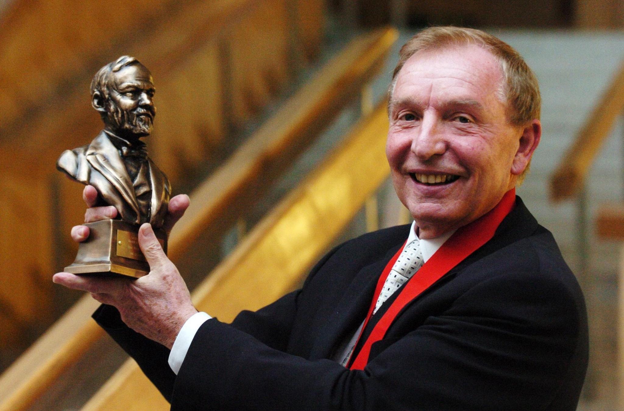 Sir Tom in dark suit and light tie holding up a bust of Andrew Carnegie. He is smiling.
