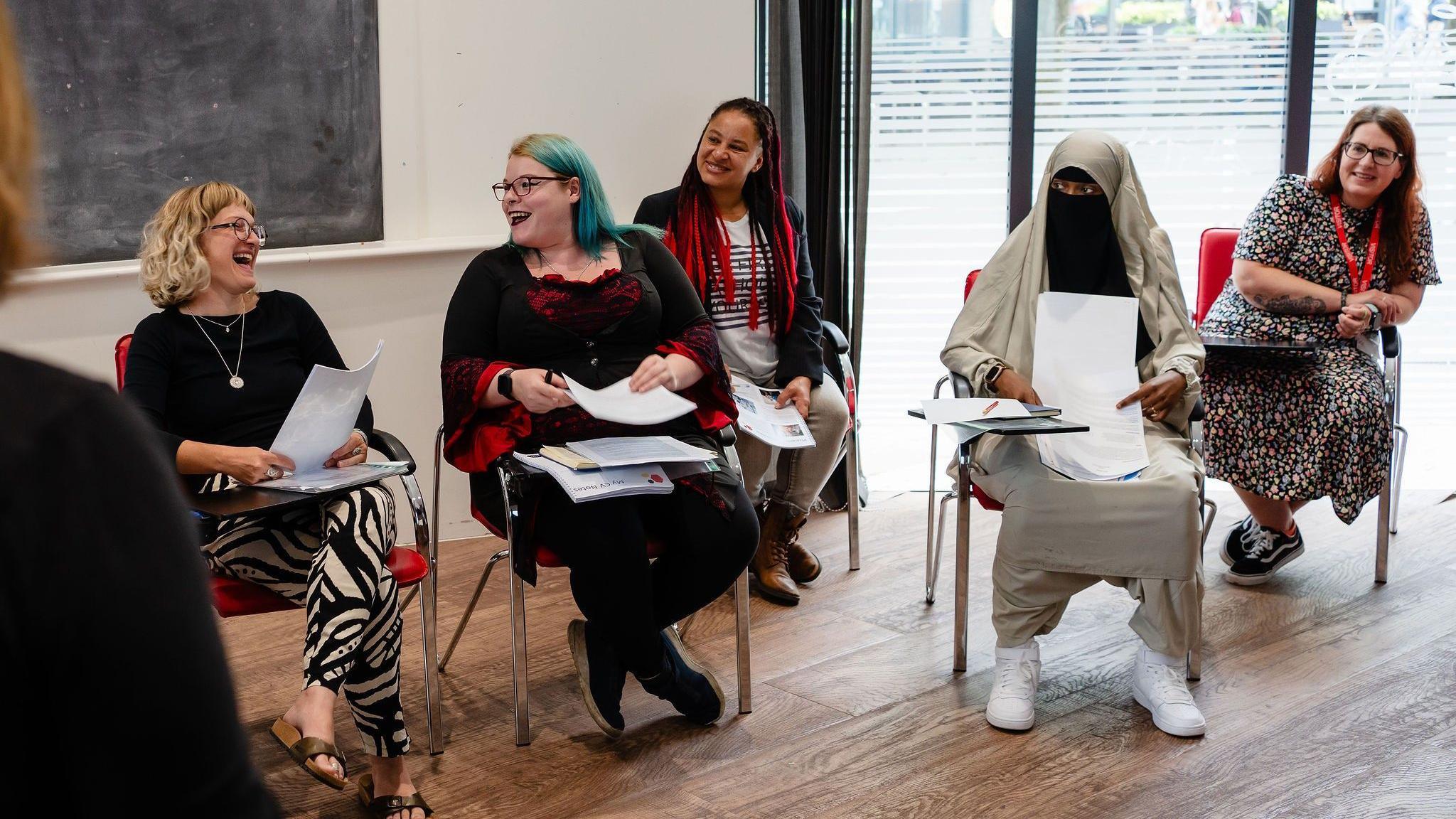 Five women are sitting in chairs with stacks of paper on their laps or on small desks attached to the arm of the chair. A blonde woman with glasses and a woman with glasses and blue hair are looking at each other and laughing. A woman with long black and red braids is smiling at them. Next to her are a woman with brown/red hair and a woman wearing a Niqab.