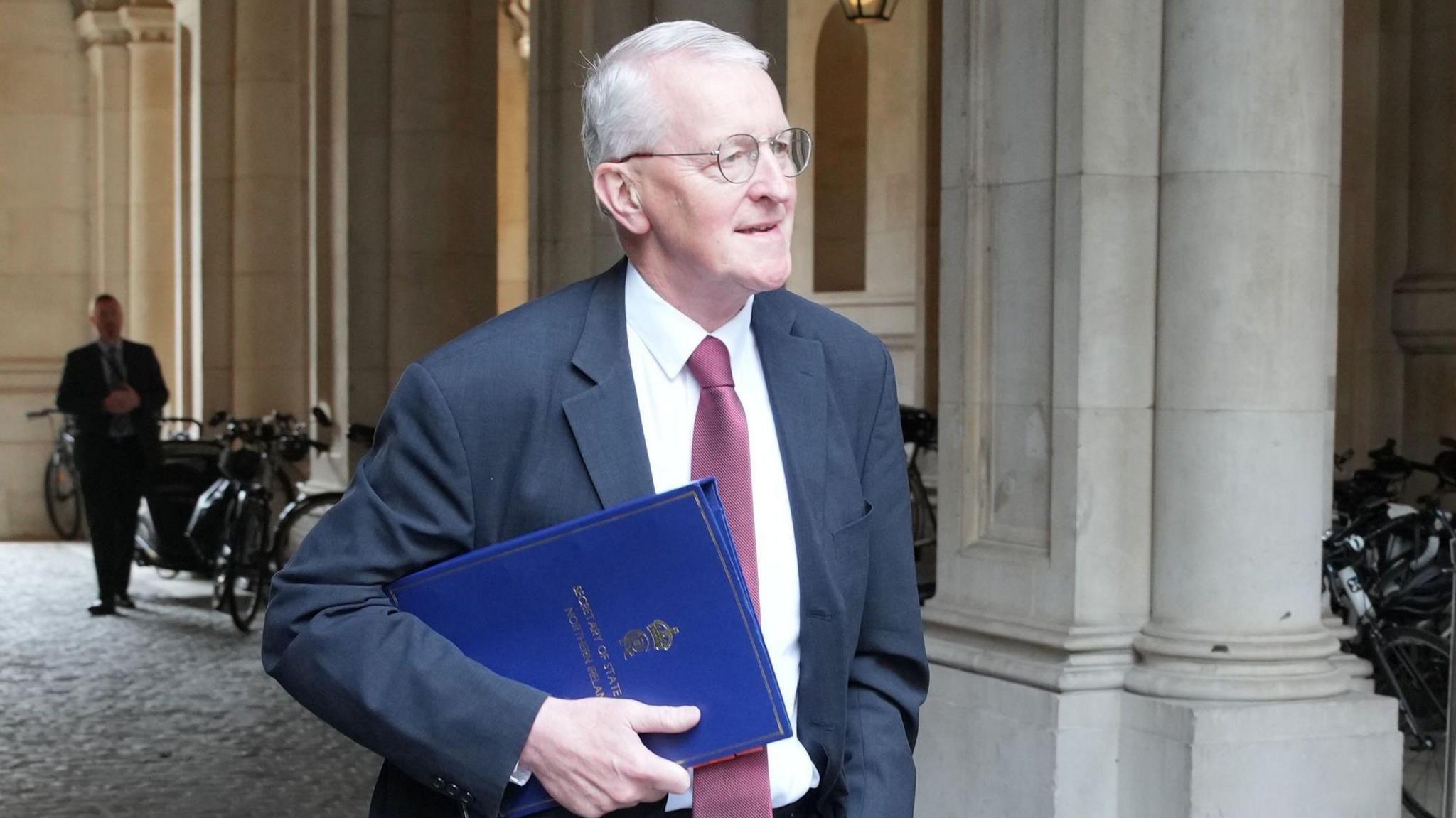 Hilary Benn wearing glasses, a navy suit, white shirt and red tie walking carrying a blue folder.