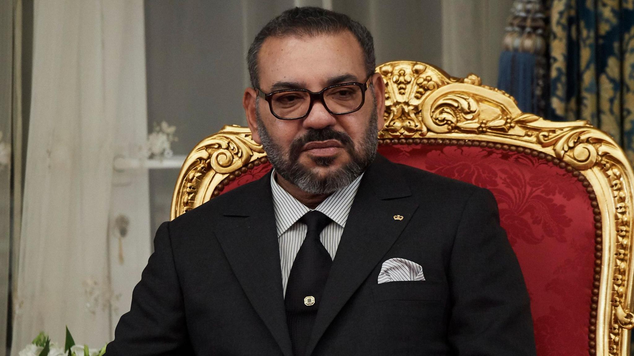 King Mohammed VI of Morocco wears a black suit and tie with matching pinstripe shirt and handkerchief in his top pocket while sitting on a throne with a red velvet seat and ornate gold surround