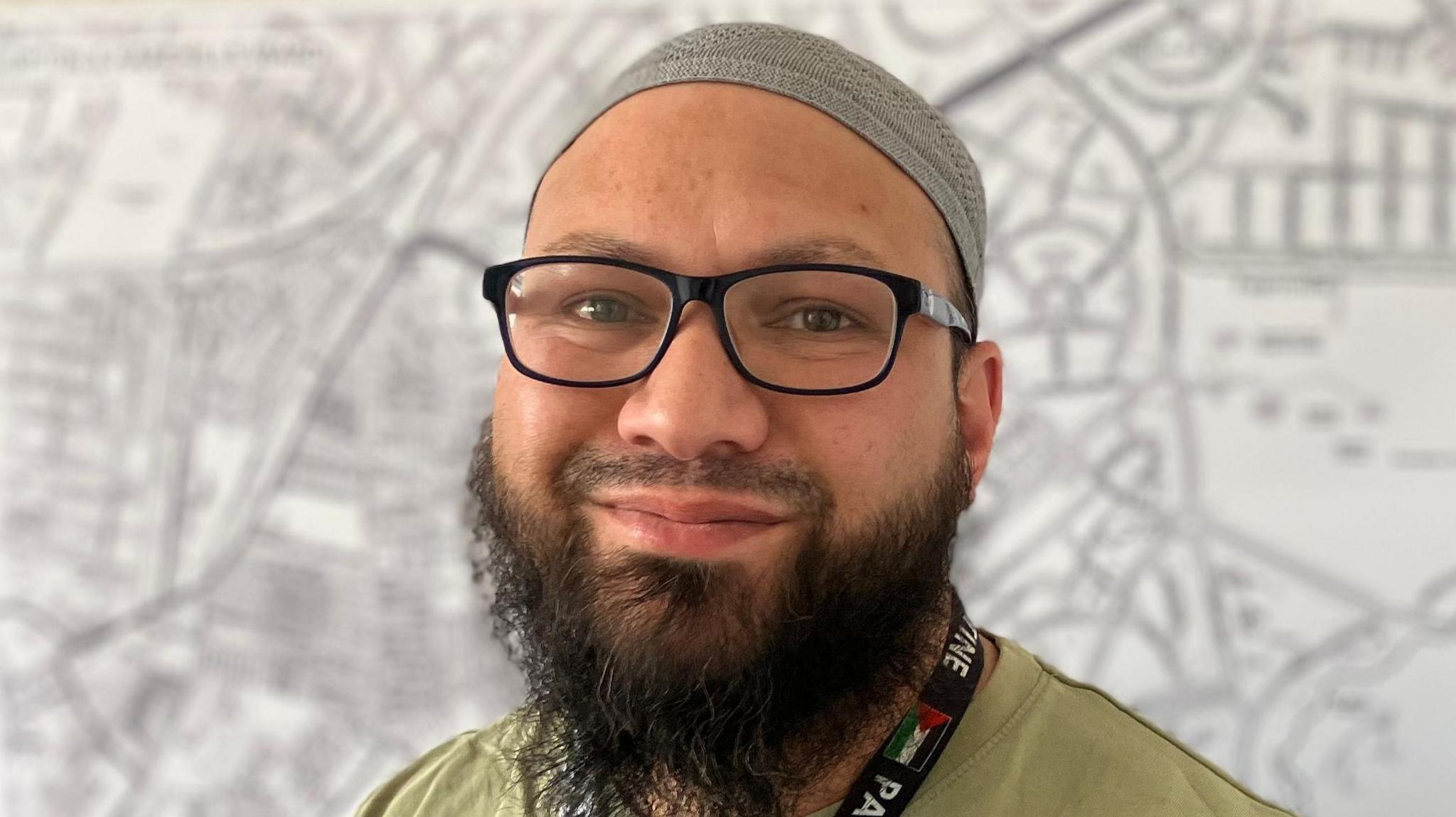 Mothin Ali, who is wearing a grey skullcap. He is wearing glasses and has a black beard. A lanyard hangs around Mr Ali's neck.
