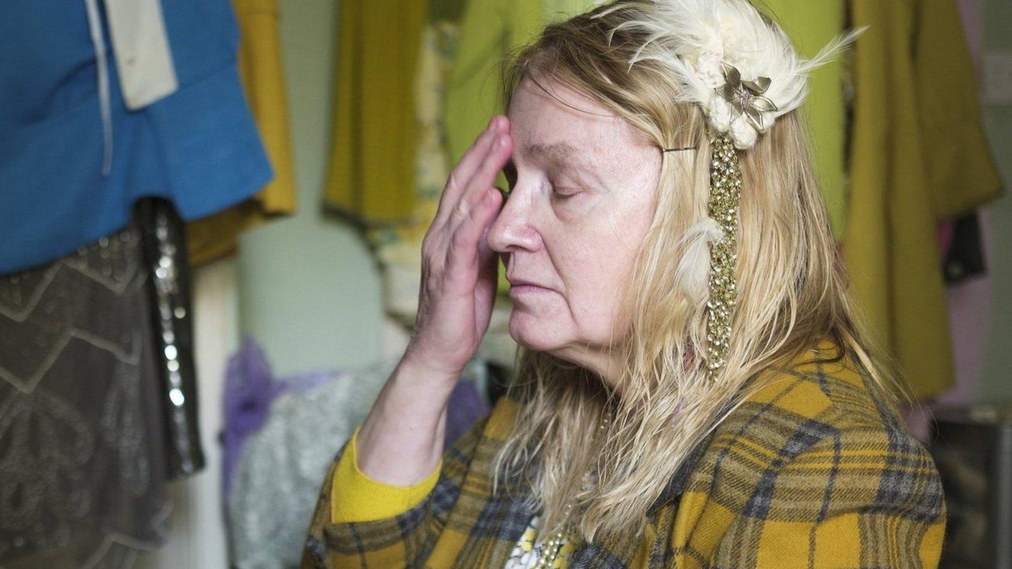 Liz holding her head sitting on her bed with dresses hanging on the walls around her