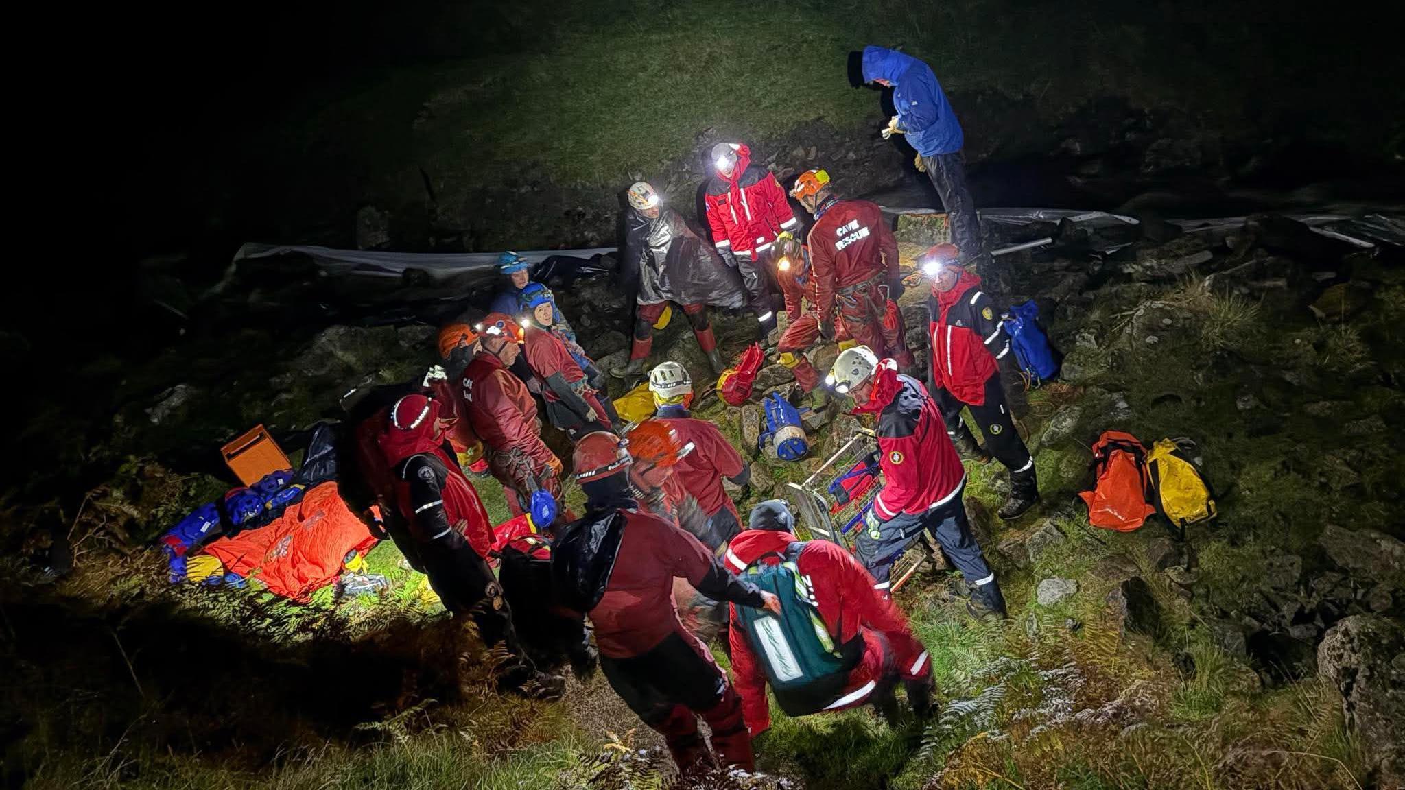 A group of individuals, dressed in red and blue jackets, helmets, and equipped with headlamps.
The scene is lit by headlamps and other portable light sources. The terrain is grassy with visible rocks, suggesting a remote or mountainous location.
One person in a blue jacket stands slightly apart from the group. Around the group, various pieces of equipment such as backpacks and bags are scattered.