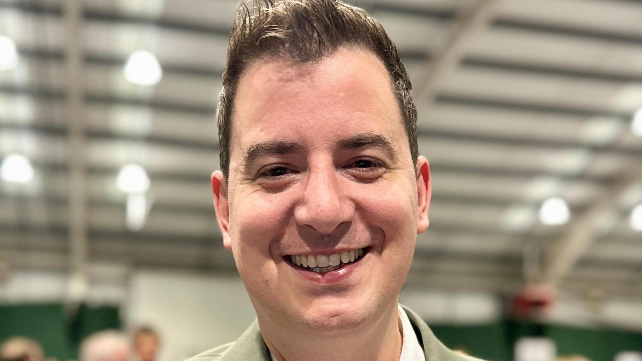 A close-up picture of Mike Reader. He is facing the camera and smiling. The large hall behind him has been blurred. He has short dark hair and he is hearing a cream jacket with a white shirt underneath.