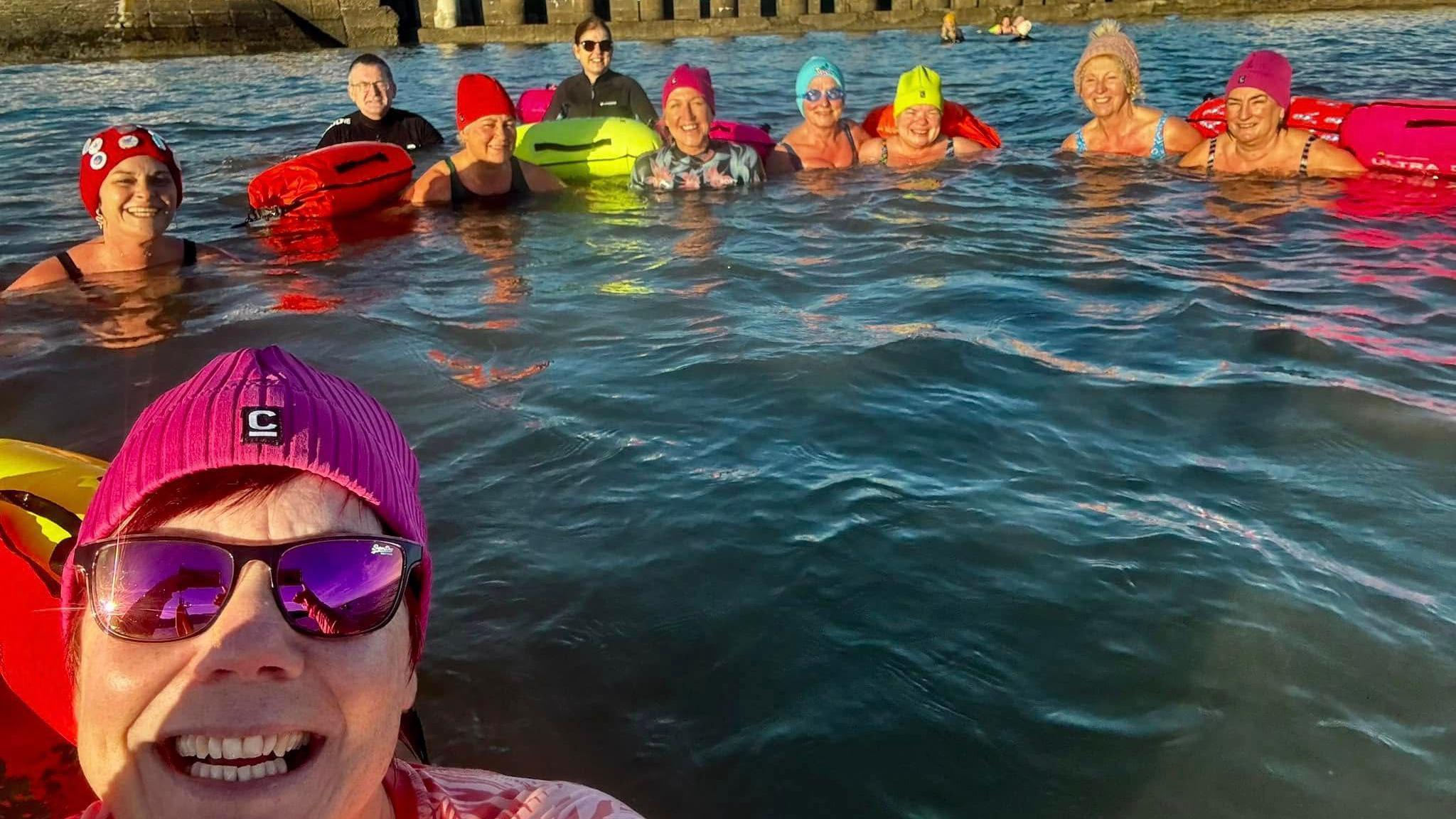 A group of swimmers in the sea, a woman in a pink and sunglasses hat takes a selfie. Nine men and women in multicoloured hats, with floats smile behind her.