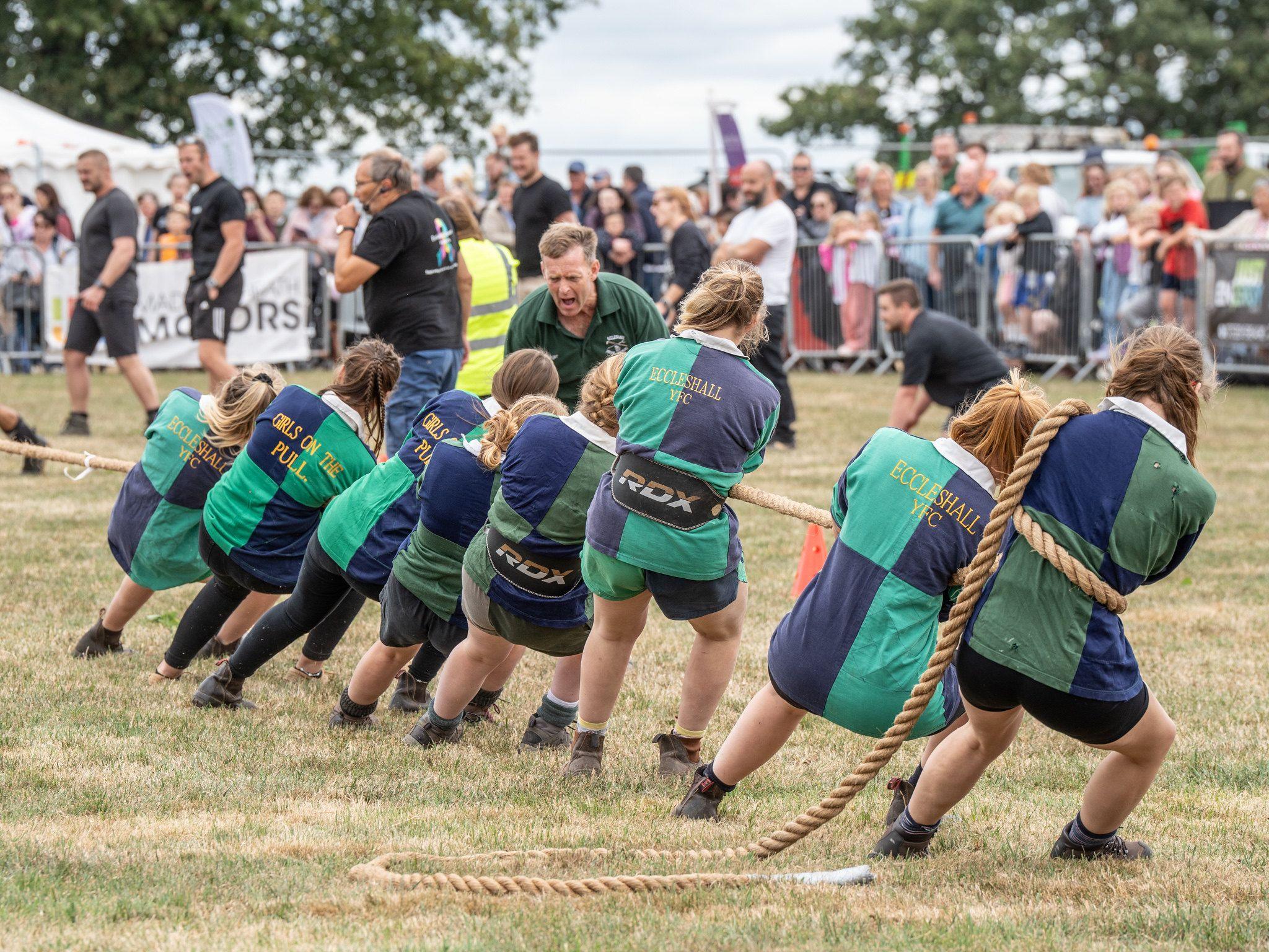 A team of eight compete in a tug of war competition. The women all wear green and blue tops and are straining to pull the rope. 