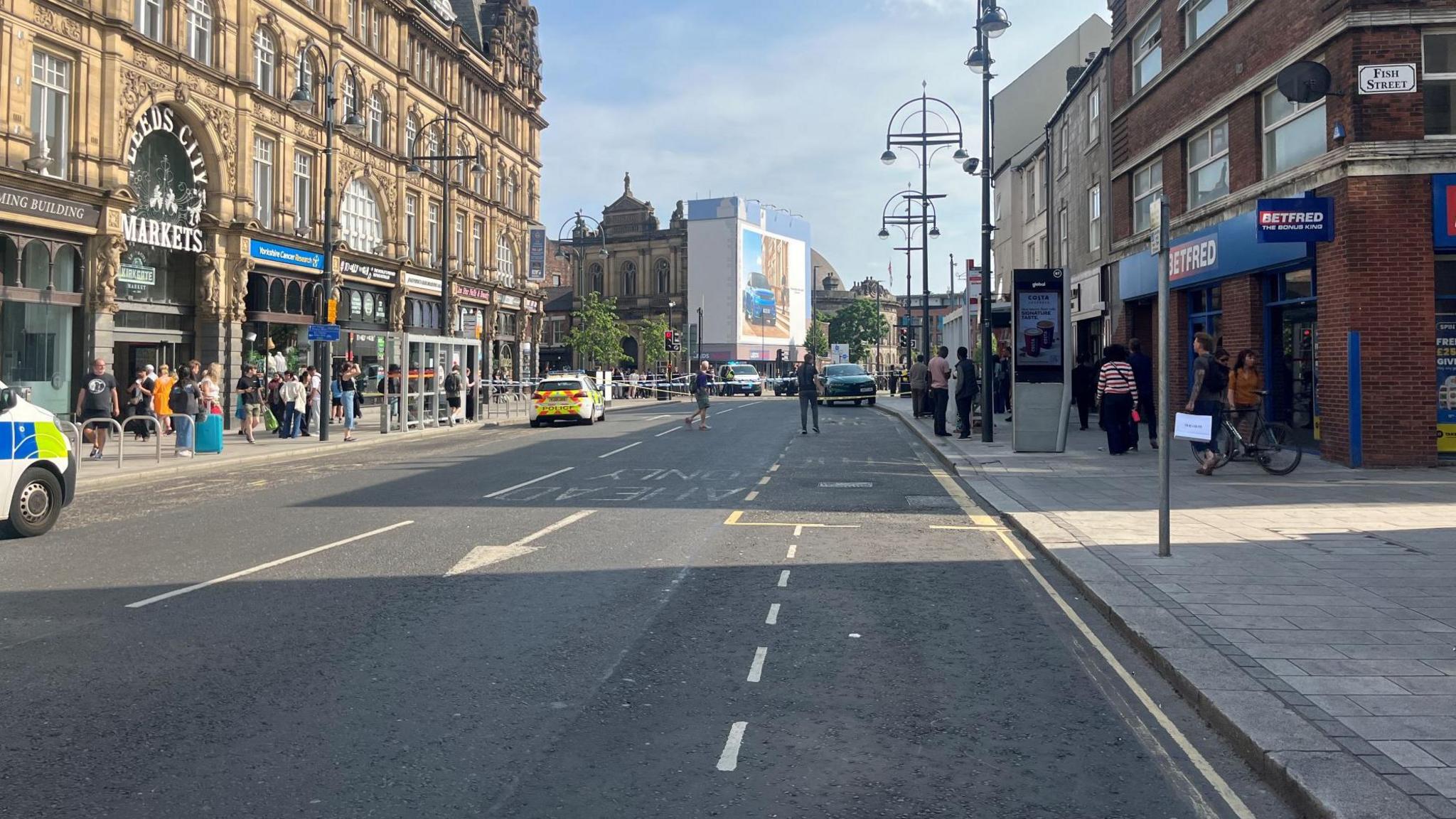 A road showing part of it with a police cordon blocking off access. On each side there are buildings and several pedestrians. There are two police cars on the road.