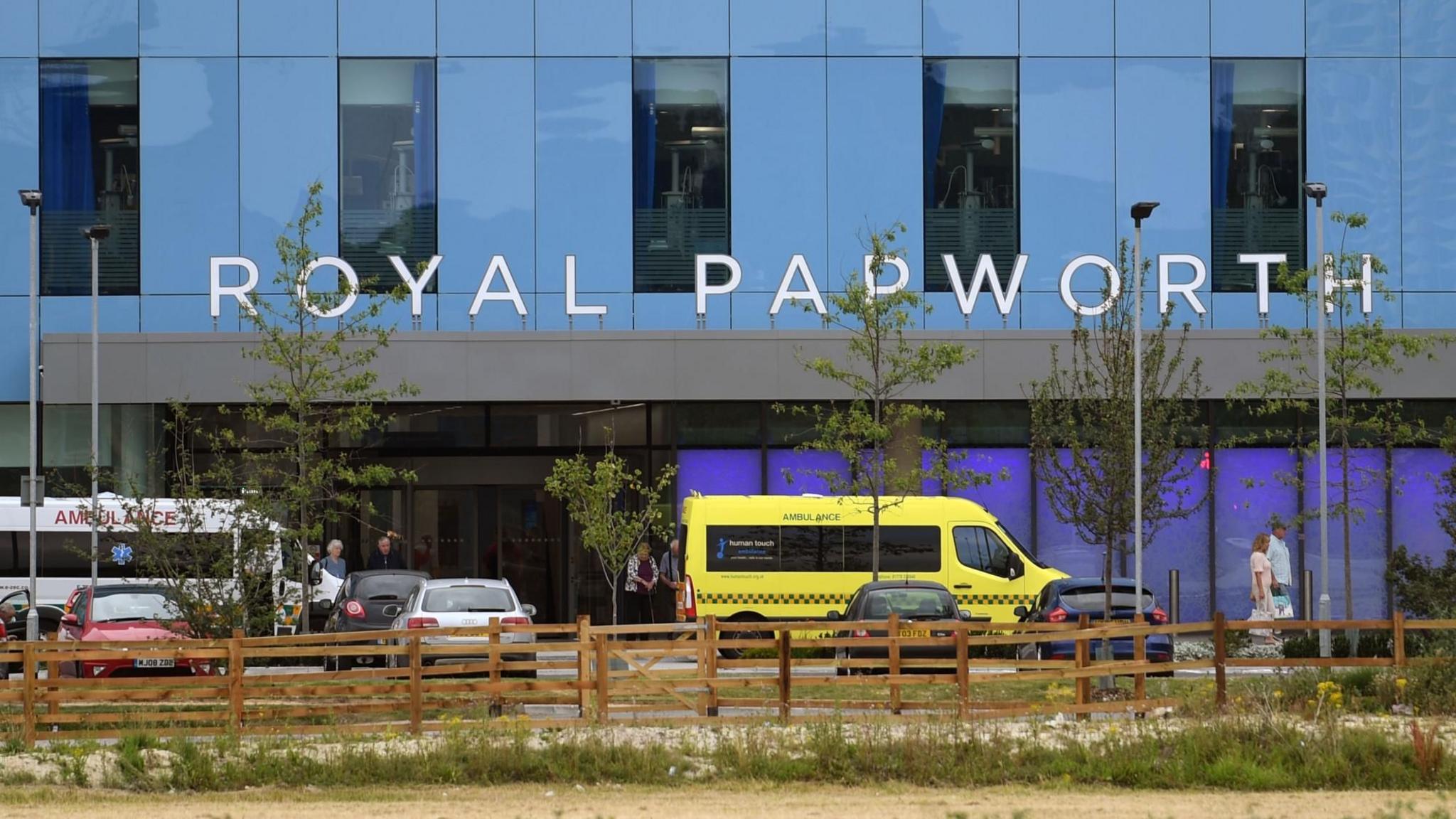 The front of Royal Papworth Hospital in Cambridgeshire. It is a new building with a shiny blue facade. The front entrance has a large grey overhang and there is a wooden fence with grass in the foreground. Two transport ambulances are parked outside the front door. One is yellow and one is white. A few cars are also parked in front of the hospital.