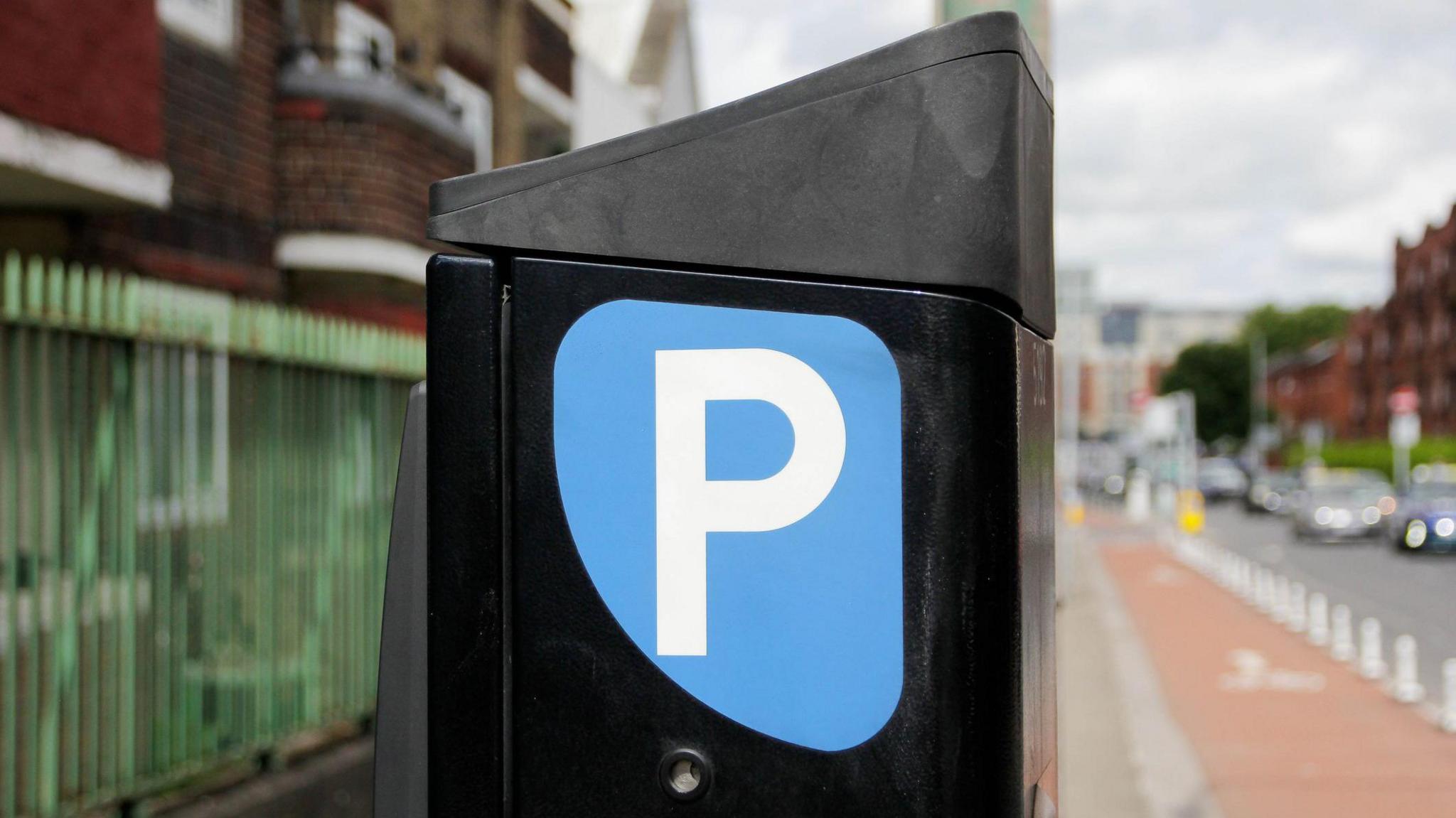 A black box with a blue bubble with the letter P in it, in white font. It sits on the black tarmac pavement with a red cycle lane running along the right hand side of it. Dark red brick flats can bee seen to its left. Cars can be seen out-of-focus driving along the road.