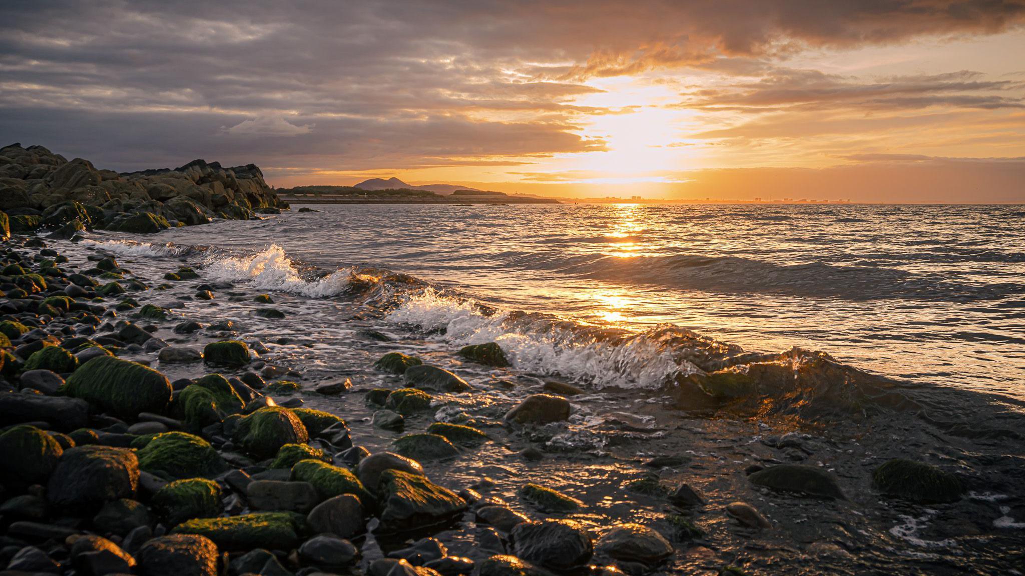 A rocky shoreline in golden sunlight. The sun, partially hidden by clouds, reflects onto the water's surface, creating reflections. Gentle waves hi against the shore.