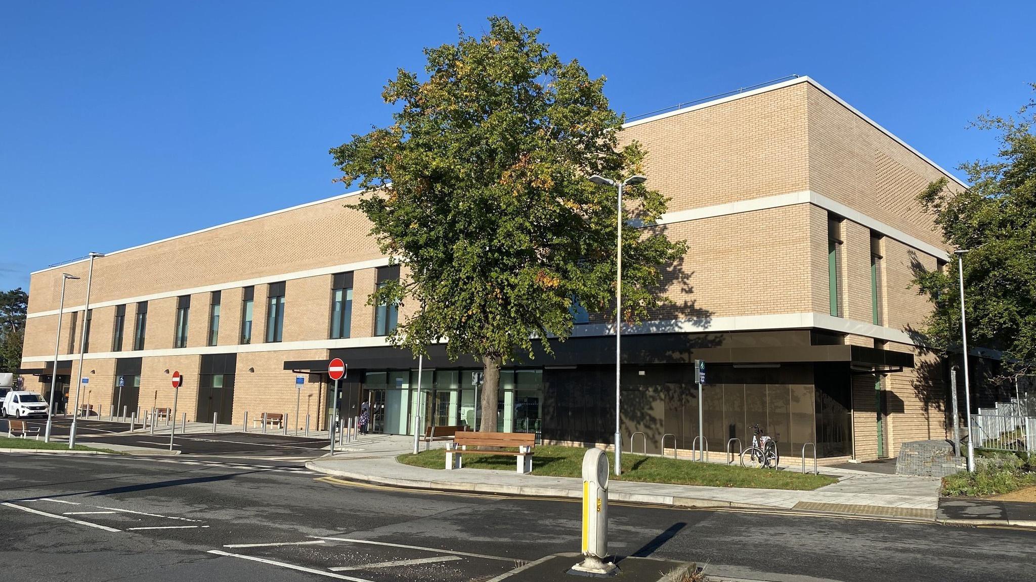 The exterior of the new surgical building. It is a large rectangular building made from sand-coloured bricks with a few vertical windows. There is a mature tree outside and some basic landscaping, with grassy patches, a wooden bench and bike rails. 