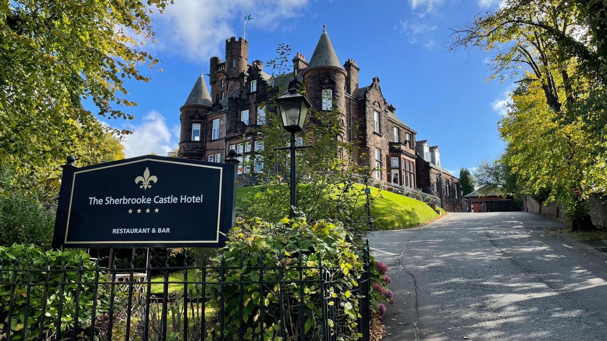 The Sherbrooke Castle Hotel in Glasgow, seen from the road. The hotel is a large sandstone building with turrets. The hotel sign is in the left of the image, among trees and bushes, and the road up to the hotel is on the right.