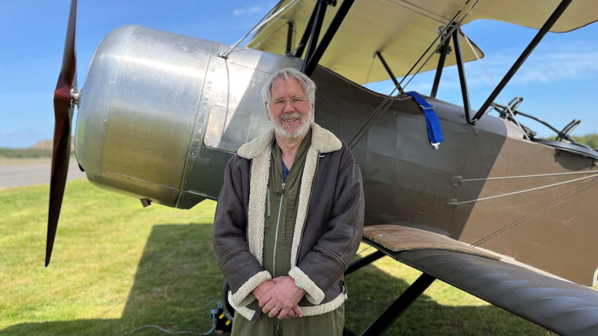 Tim Rayner, a white-haired man with a beard and glasses, stands in front of the plane, wearing a khaki flightsuit and a leather and sheepskin flying jacket. The plane's propeller is on the left of the picture.