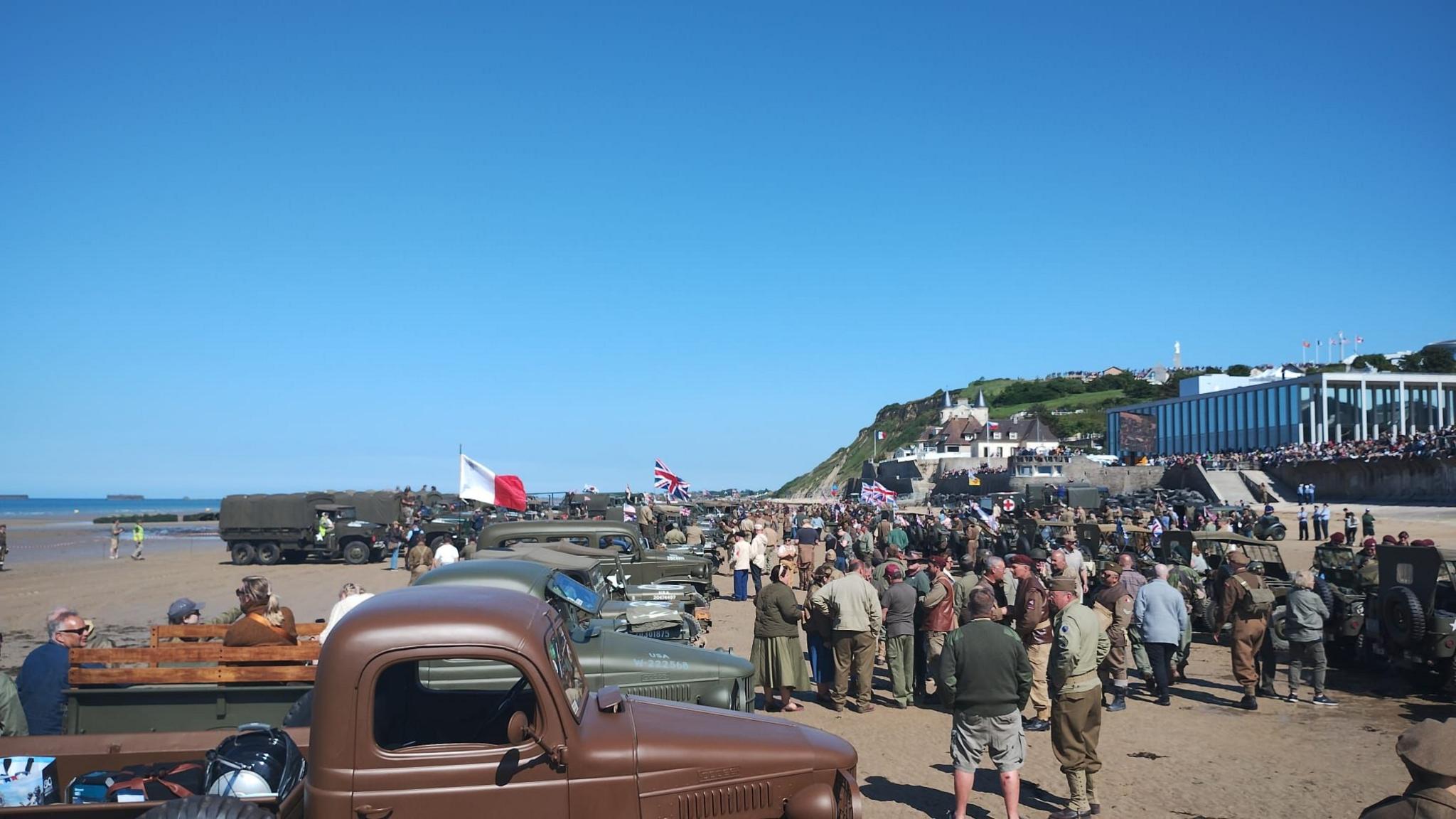 A crowd on a beach in front of vintage vehicles with blue sky