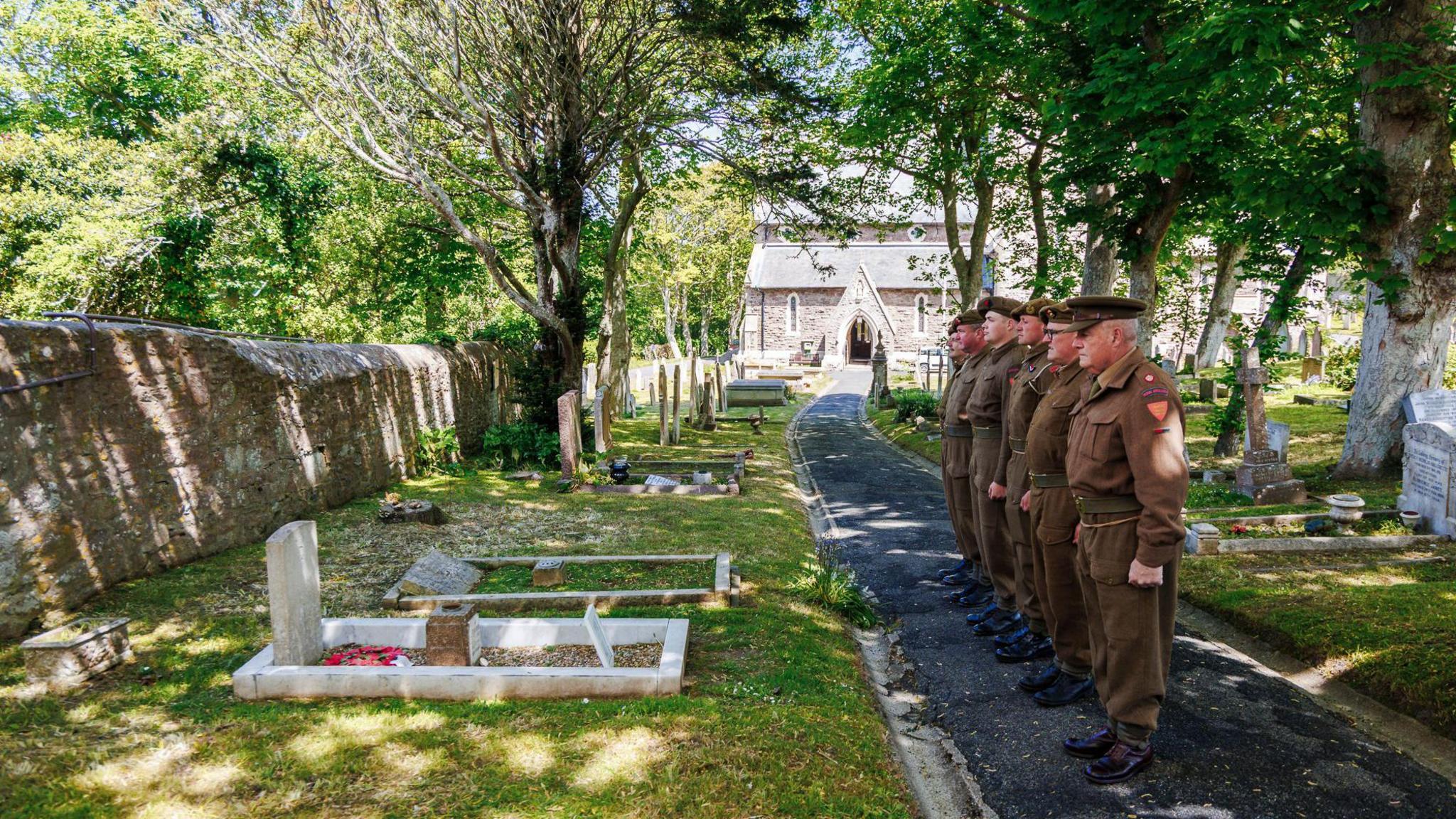 The image shows a group of six soldiers standing in a line on a paved path within a cemetery. They are dressed in historical military uniforms from the World War II era, with brown tunics, trousers, and caps.