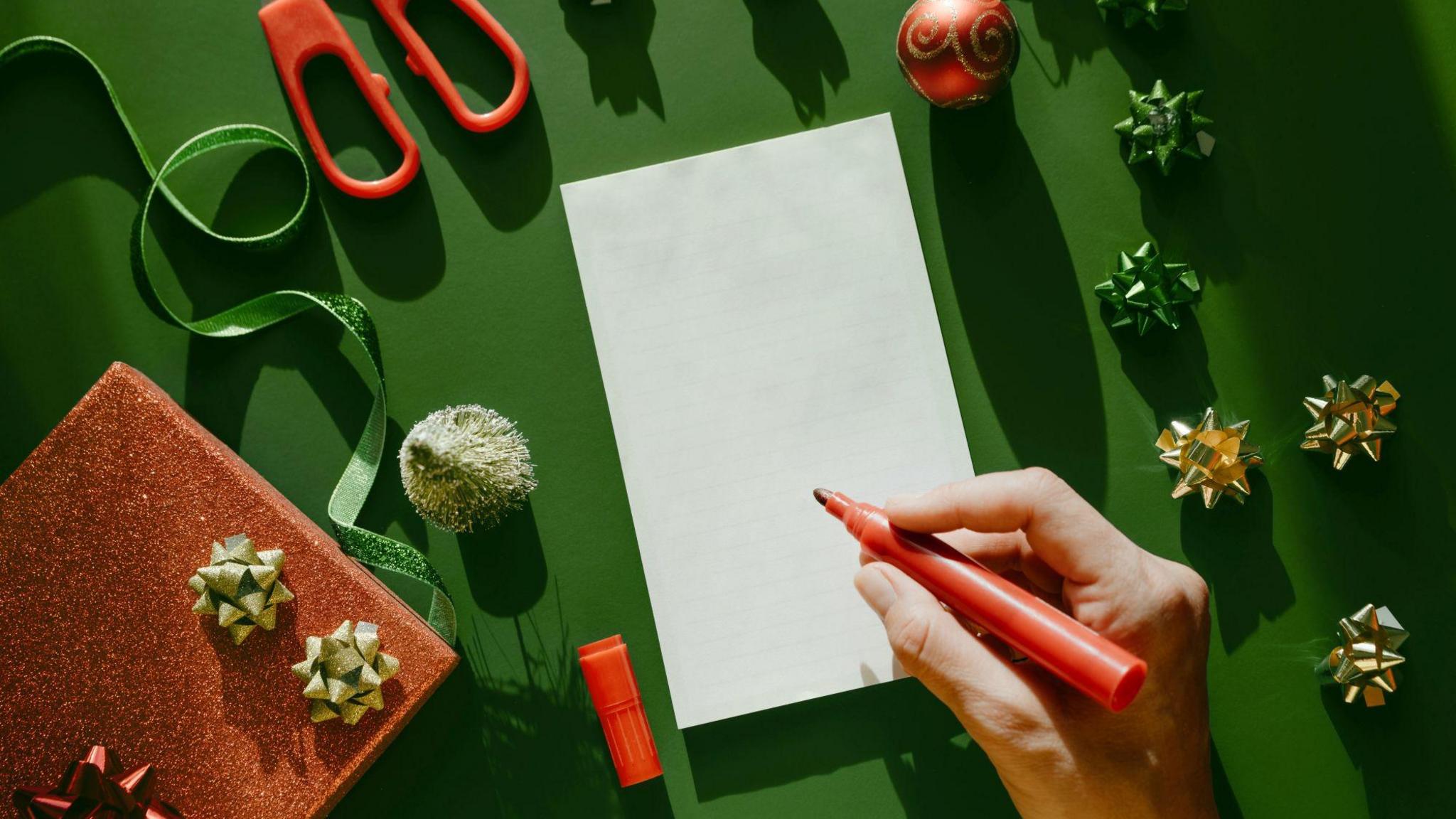 A stock image of a pad of white paper against a green background. A person is holding a red pen. Surrounding the paper are gold and green present bows, a red glitter box and a pair of scissors with red handles.