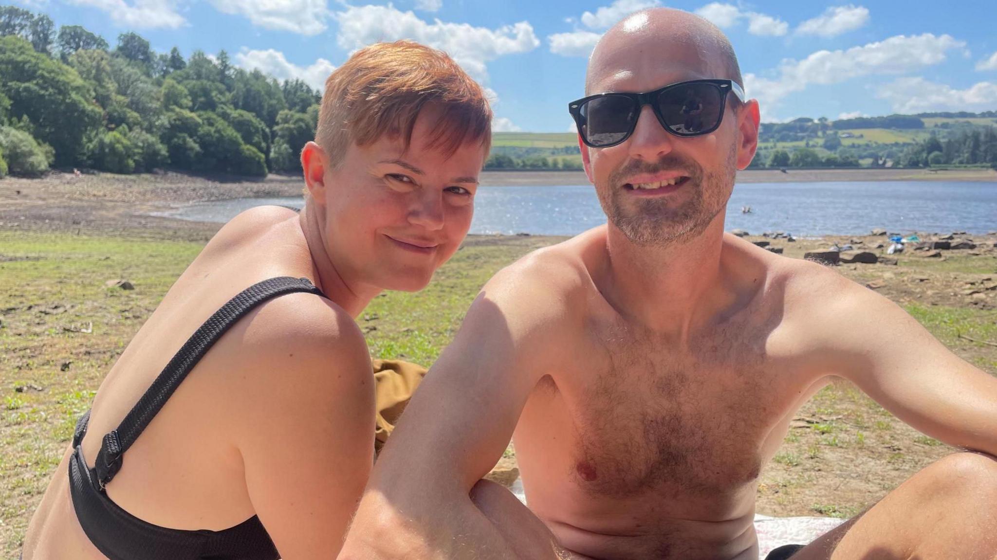 A young man and a woman in swimming costumes pose on beach towels next to the reservoir