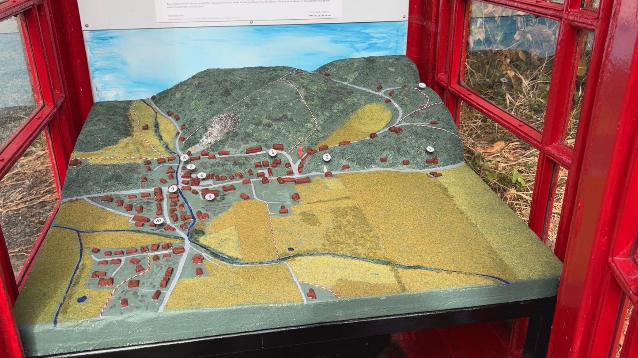 A 3D model of the village and its surrounding roads, fields and hill are displayed inside an old red BT telephone box. On the model are tiny brown buildings. Ten of them are numbered.