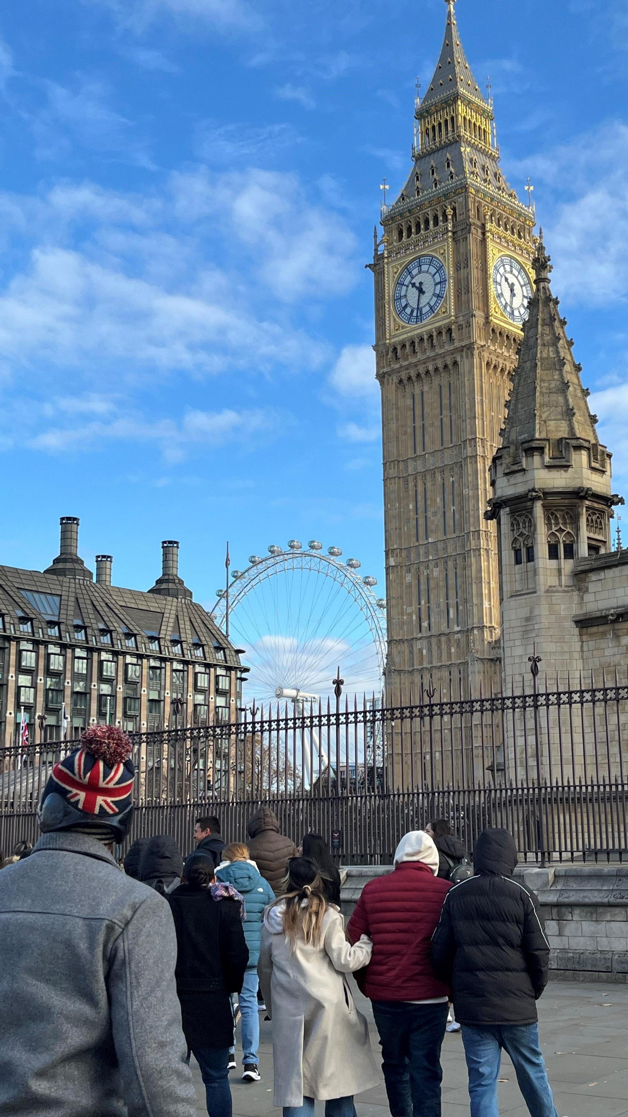 Tourists, including one wearing a union jack hat, crossing the road towards Big Ben, with the London Eye visible in the distance