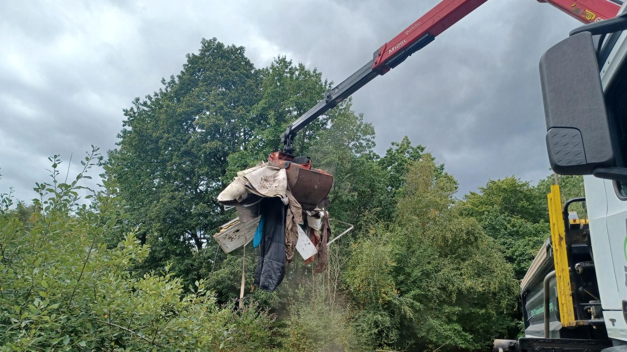 A crane carrying a load of rubbish to the back of a truck in woodland