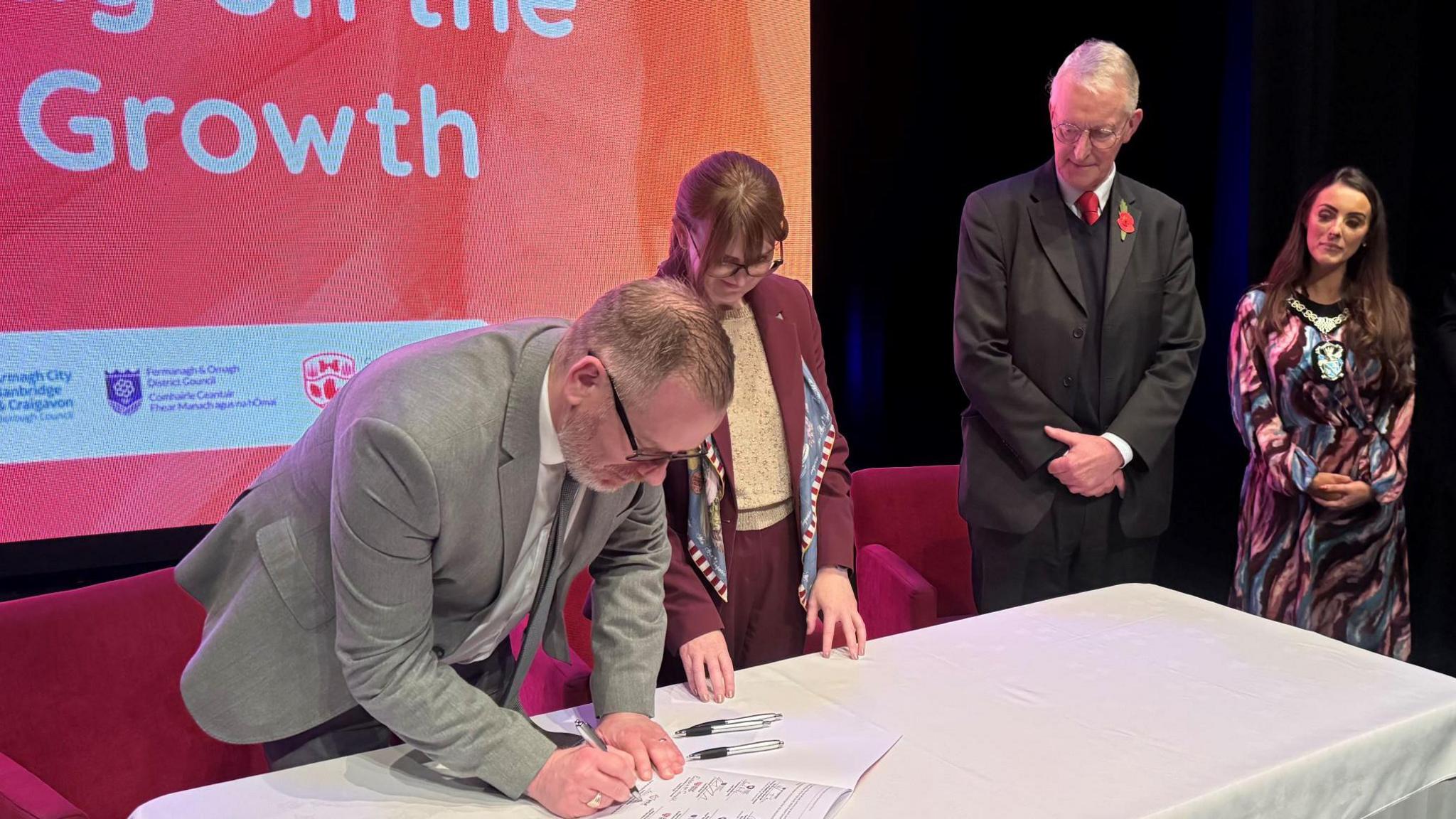 A man with short brown hair, glasses and a white beard signs a sheet of paper which lies on a white cloth table. To his right stand two women and a man.