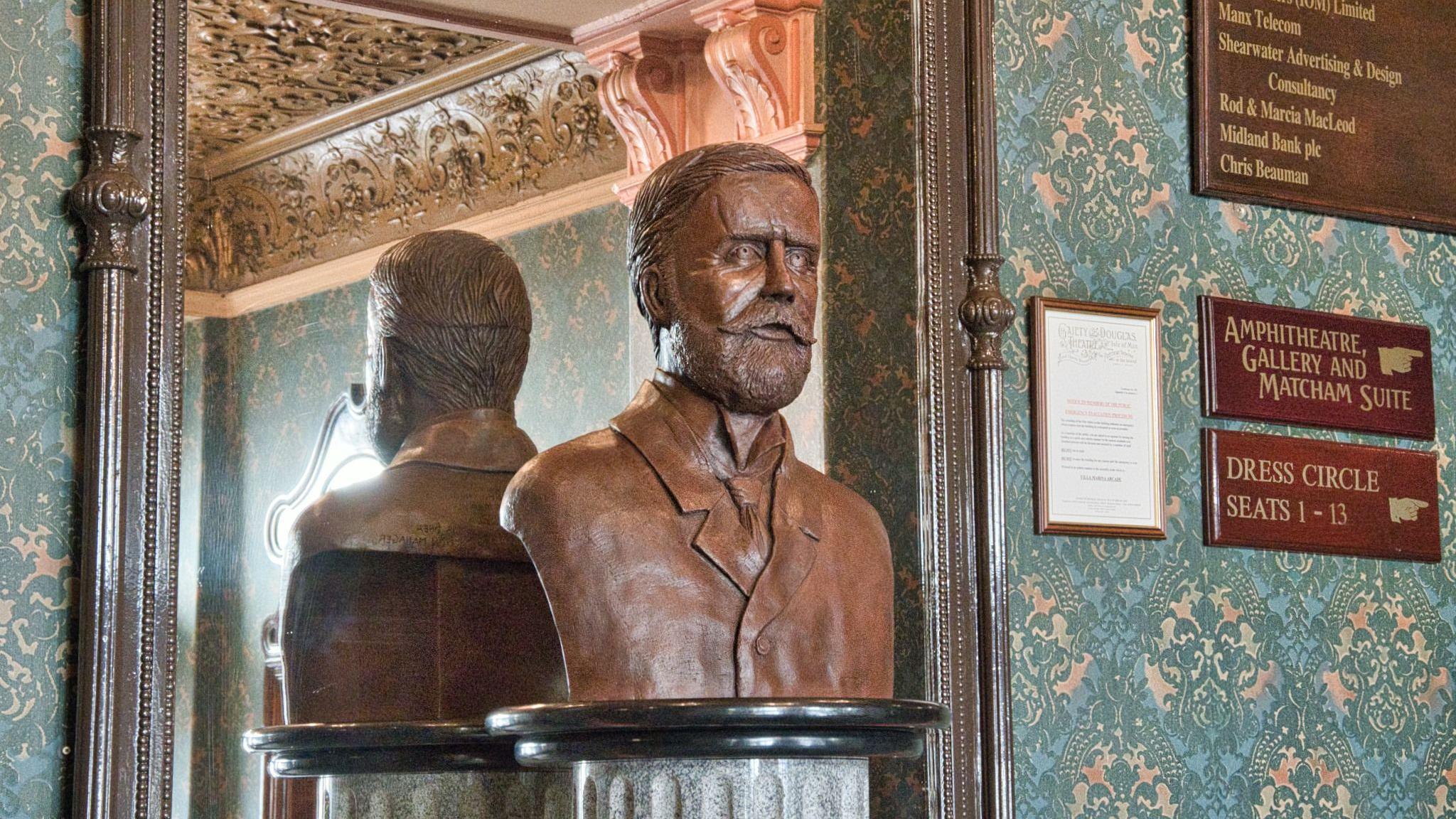 A bronze bust of Frank Matcham, which has short hair, a moustache and beard and is dressed in a jacket and tie. There is a large wooden framed mirror behind the bust which is showing a reflection of its back. The wall is covered in highly decorated green coloured wallpaper and there are wooden signs pointing to the seating areas on the right.
