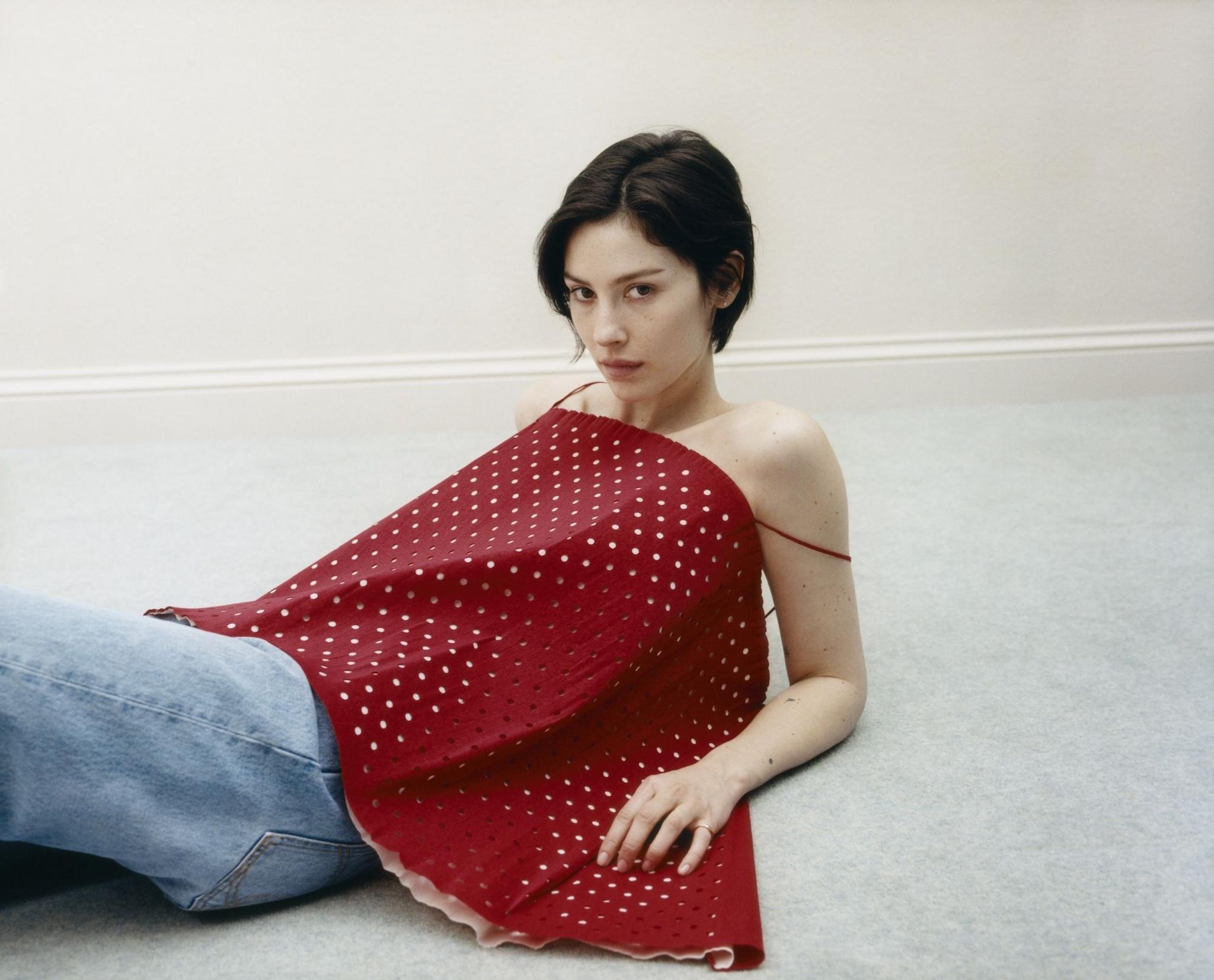 Gracie Abrams in a promotion photo for her third album, Daughter from Hell. She is lying down, propped up on her elbows, in a plain room; wearing a strappy polka dotted top.