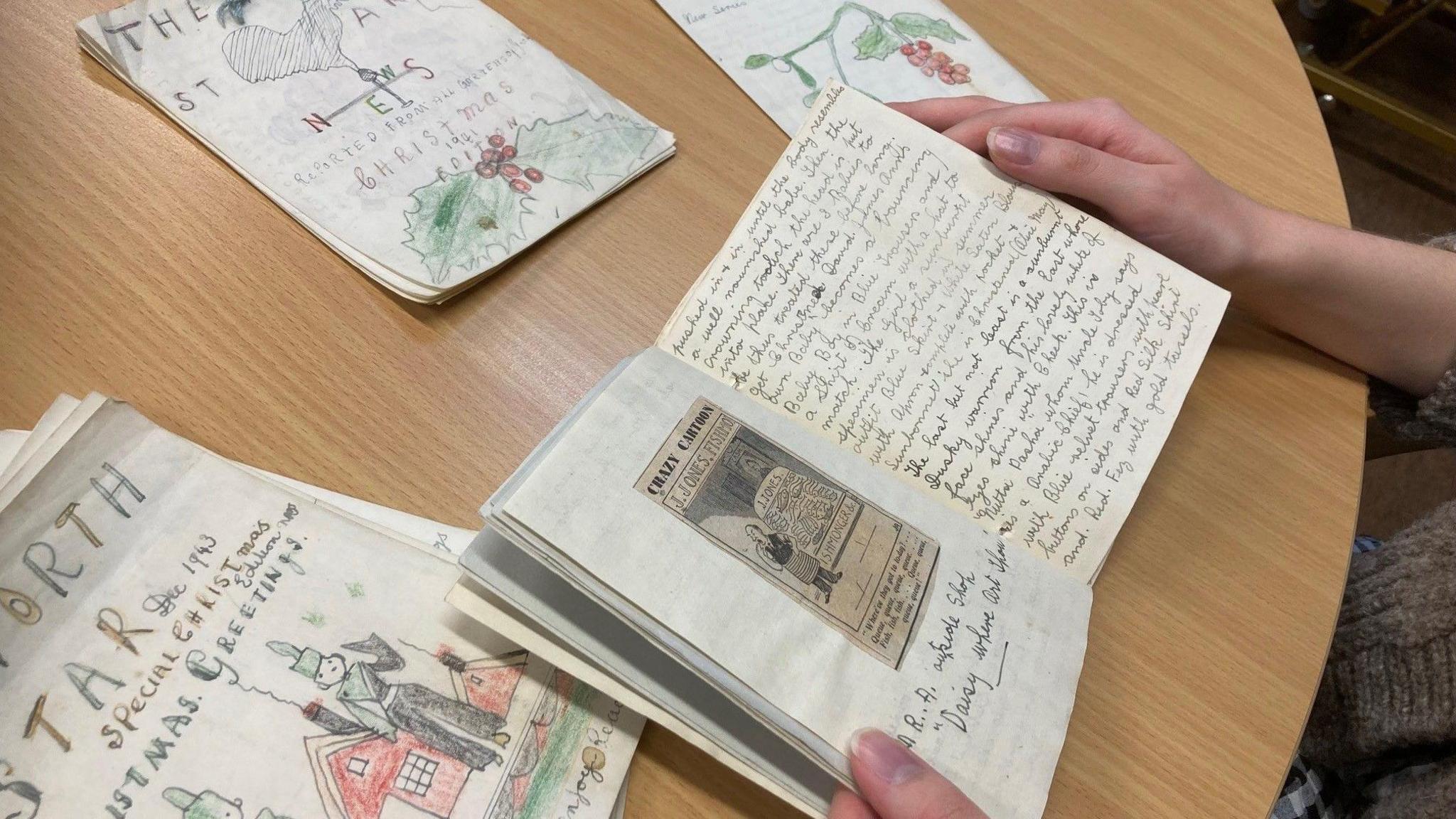 A close-up of hands holding the pages of a small paper book, handwritten in pencil and hand-illustrated, over a wooden table, with others lying alongside it.