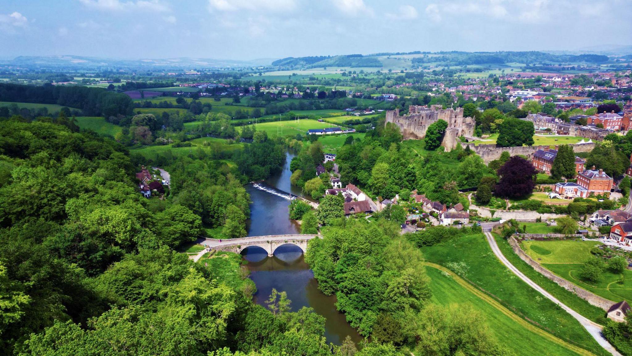 An aerial drone shot looking over Ludlow. You can see the River Severn with an arched bridge going over it. To its right is Ludlow Castle, with green ;and and houses, and the town in the distance. In the far background are more houses and rolling green hills and fields