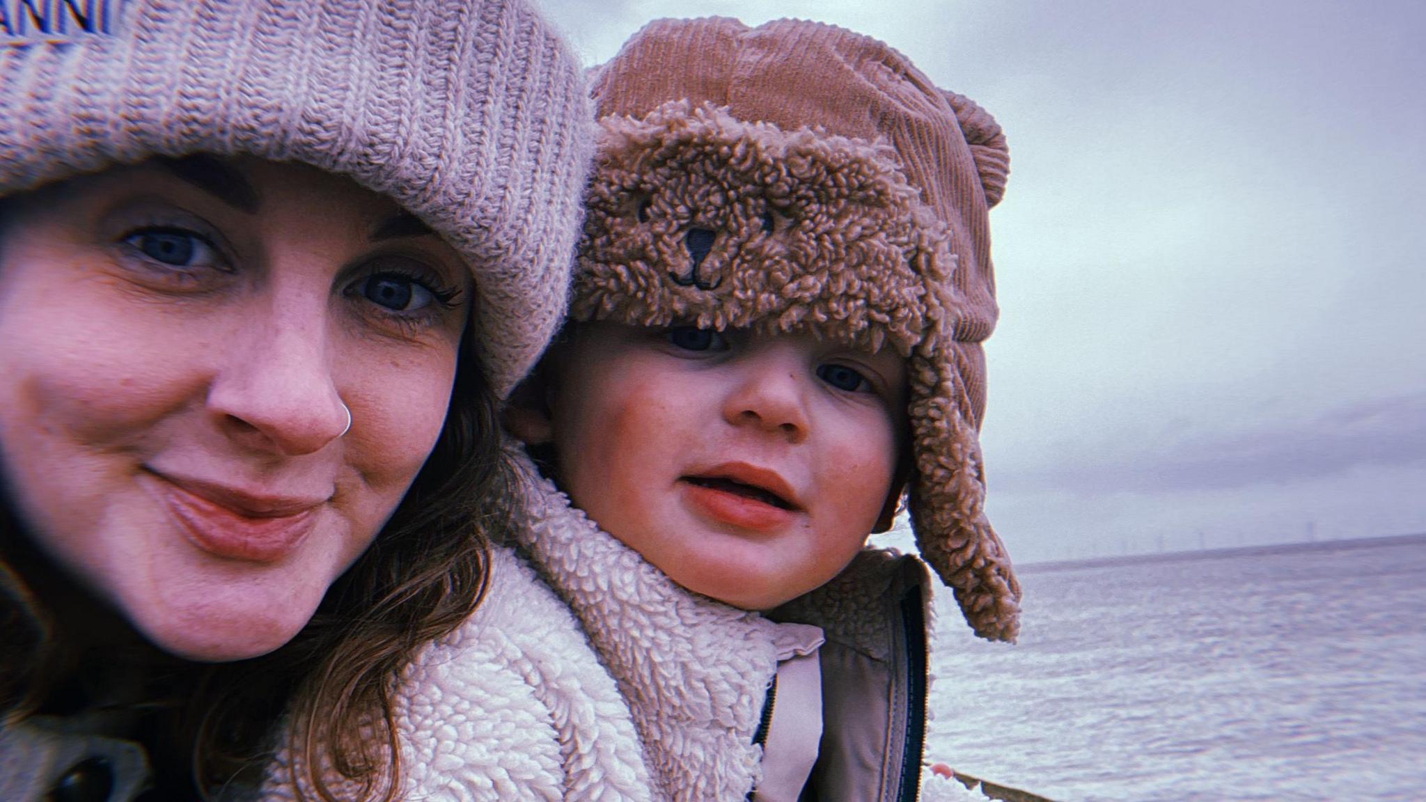 Fran Barrett stands on New Brighton promenade with her son Eli . They are both  wrapped up warm and are wearing hats as they stand in front of the sea.