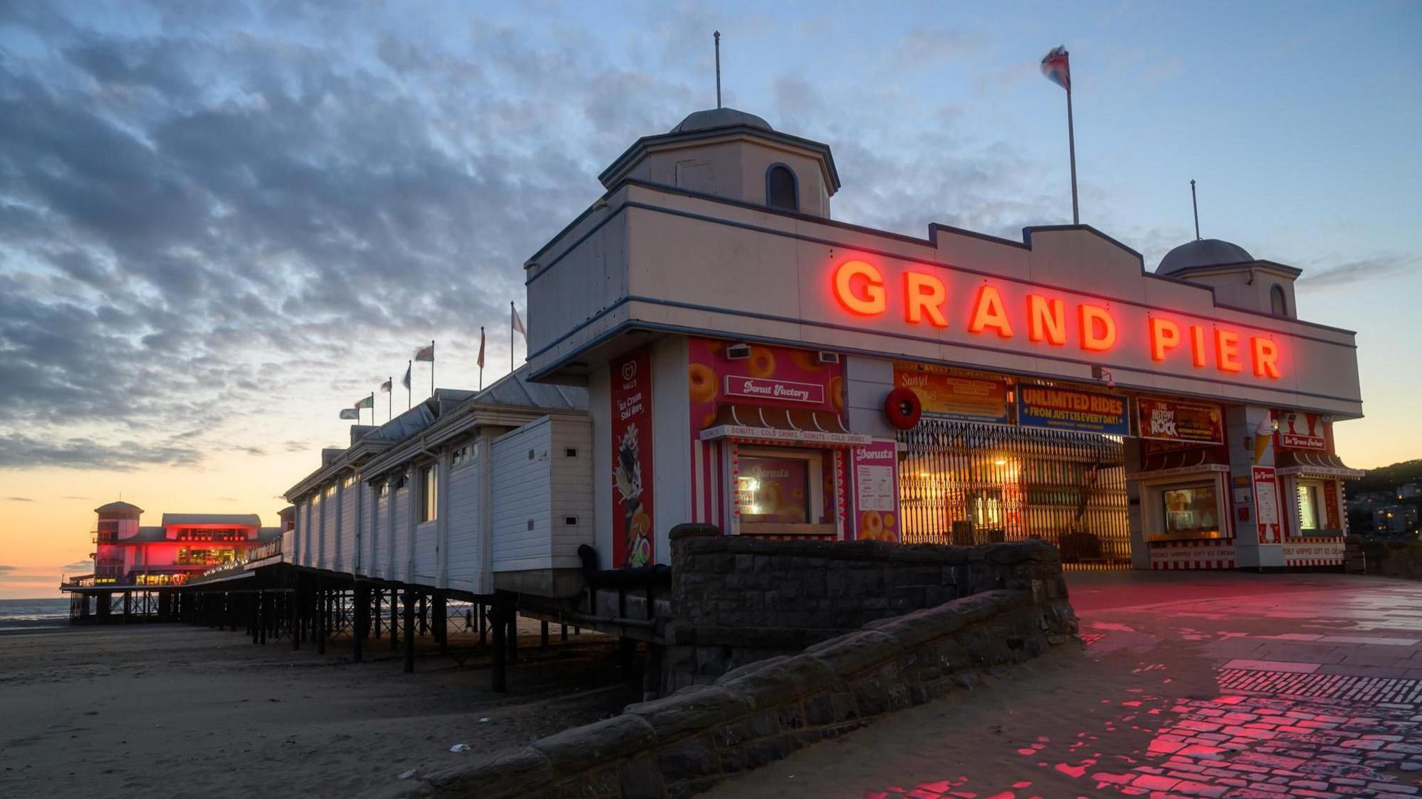 Weston-super-Mare pier is pictured at sundown. At the entrance the words 'Grand Pier' stand out in almost a neon orange/red colour. That light is reflected on the cobbles in front of the entrance. The beach and sea can also be seen and there are dark clouds passing overhead.