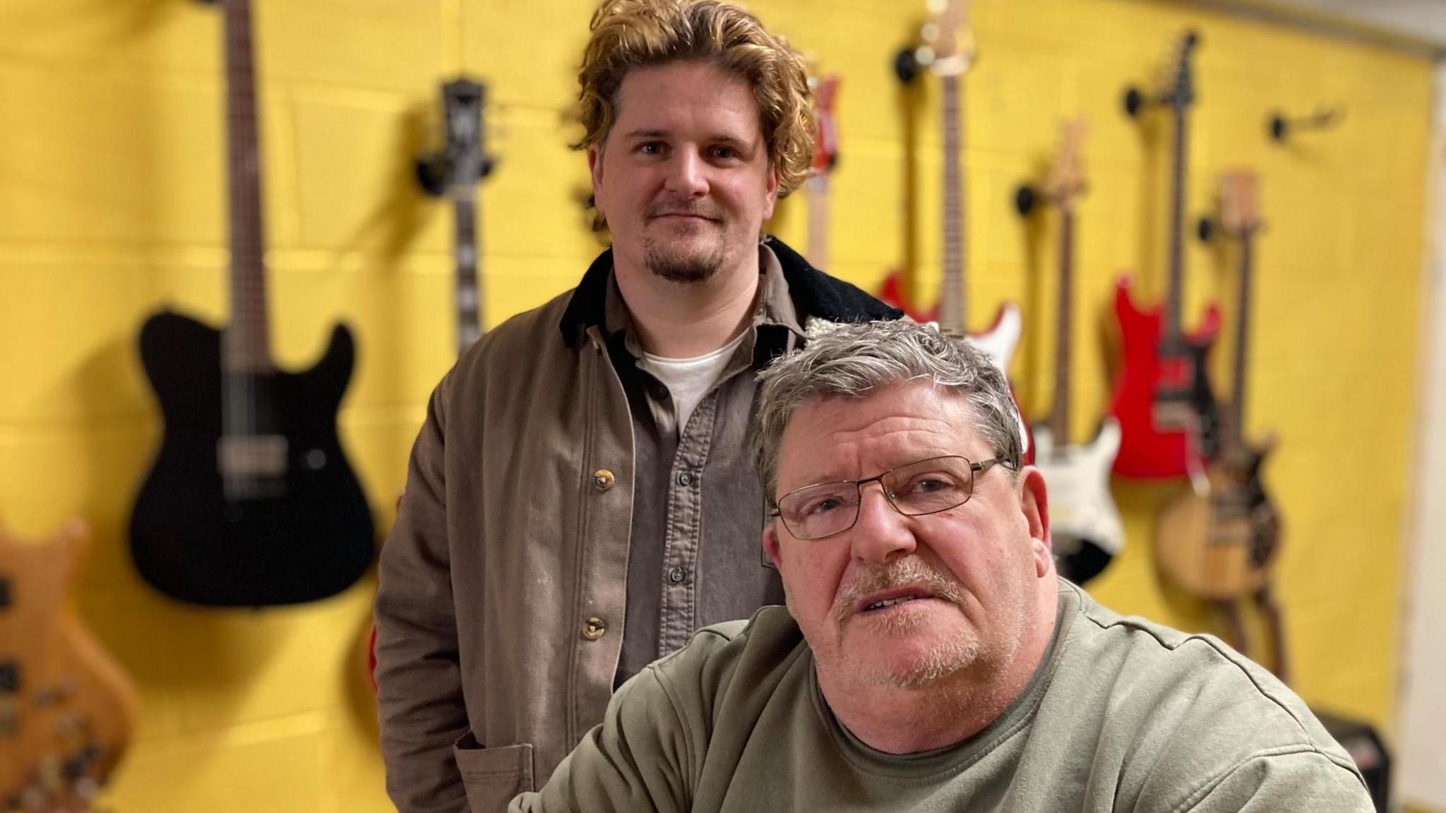 An old heavy set man with a goatee beard sits infront of a younger man who stands behind him with curly hair. A row of guitars are lined up on the wall behind.