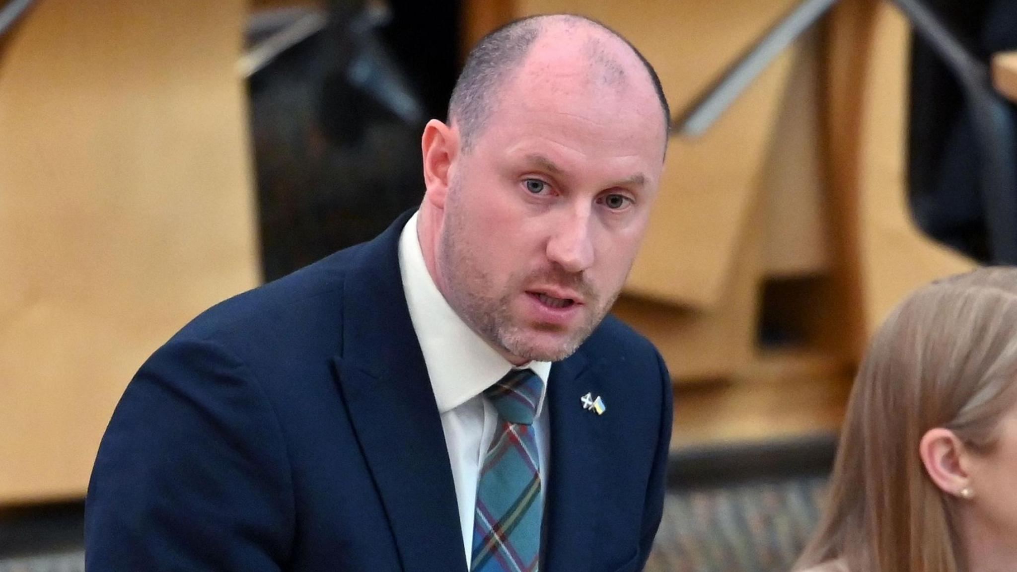 Neil Gray is taking part in a debate in the Scottish Parliament. He is wearing a dark blue suit jacket and a blue, green and purple tartan tie.
